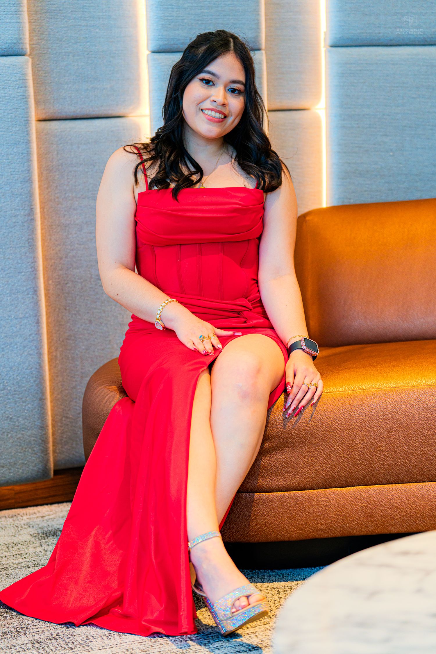 Elegant woman in a red slit gown with silver heels seated gracefully on a leather sofa indoors.