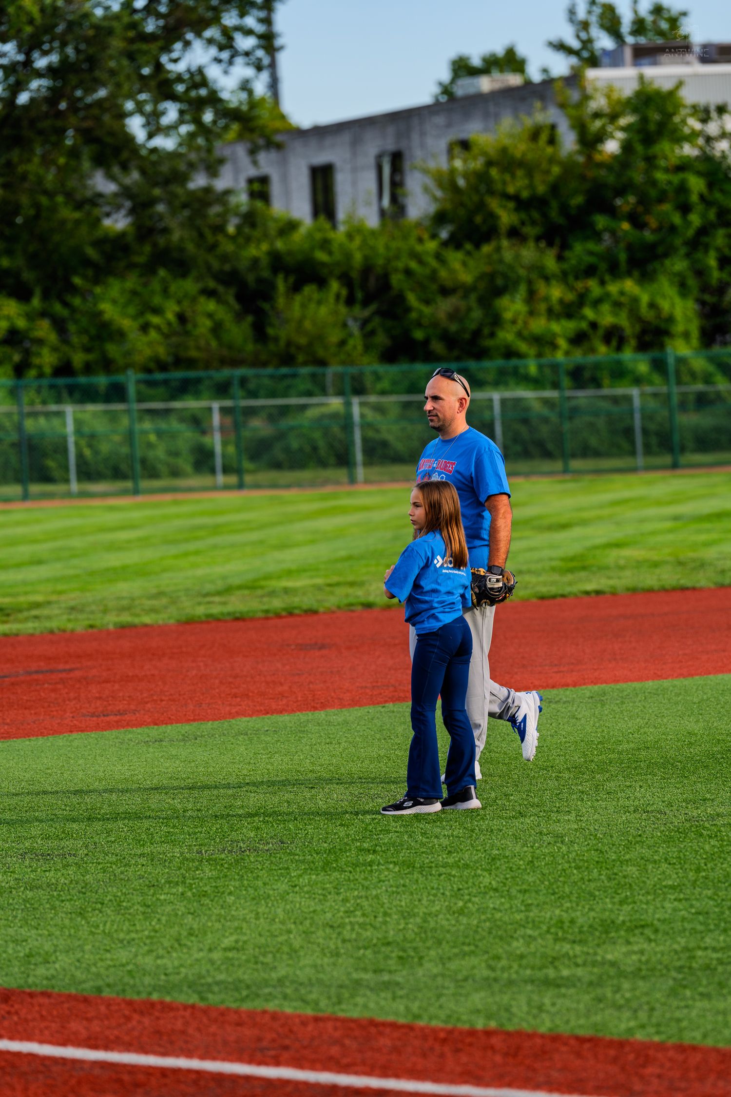 Adult coach in blue shirt standing with young girl on red and green baseball field.
