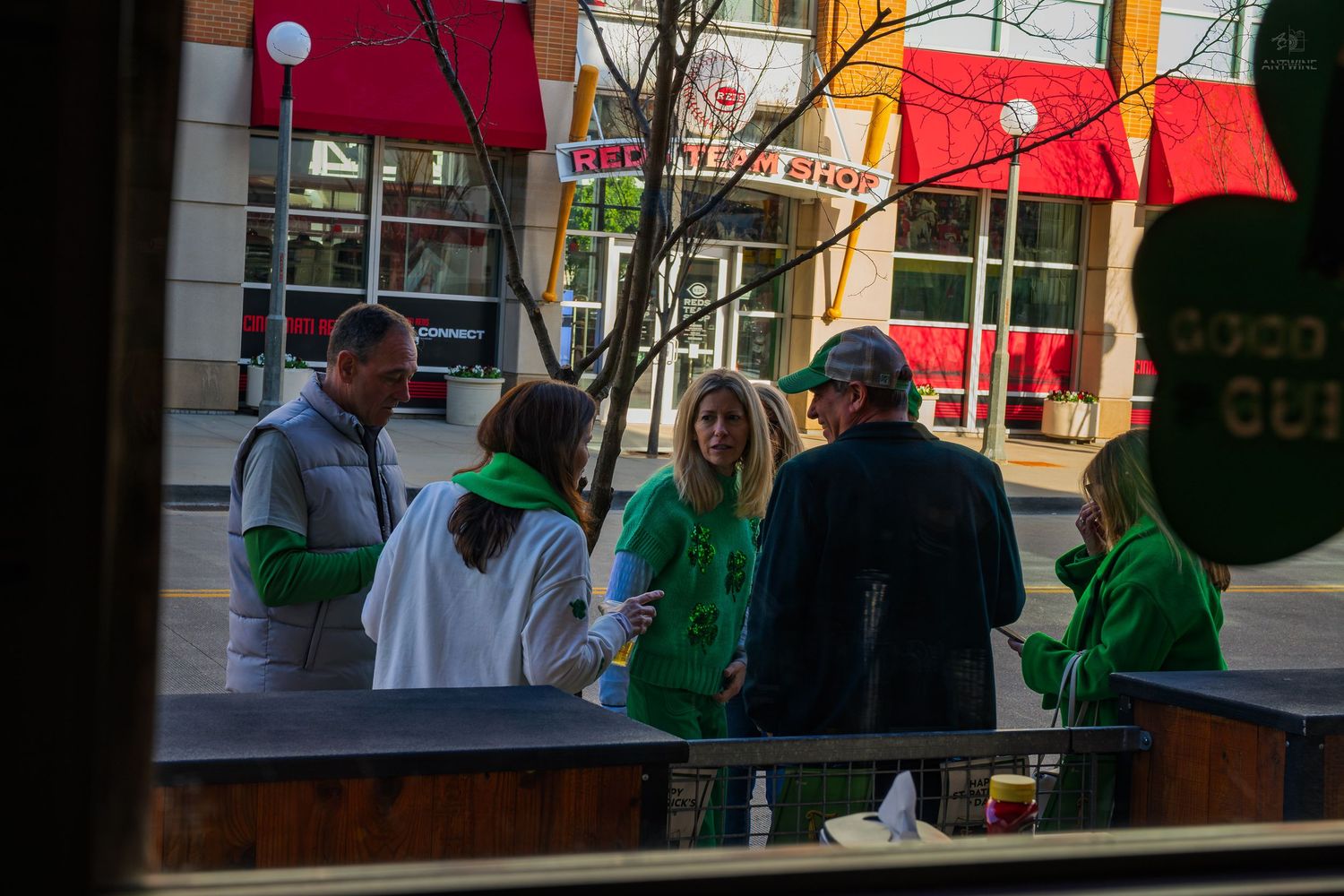 Group of people in green shirts gathered outside a urban street cafe on a sunny day.