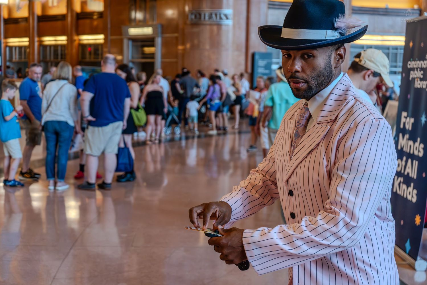Stylish man in pinstripe suit and fedora hat using phone in busy indoor venue with crowd in background.