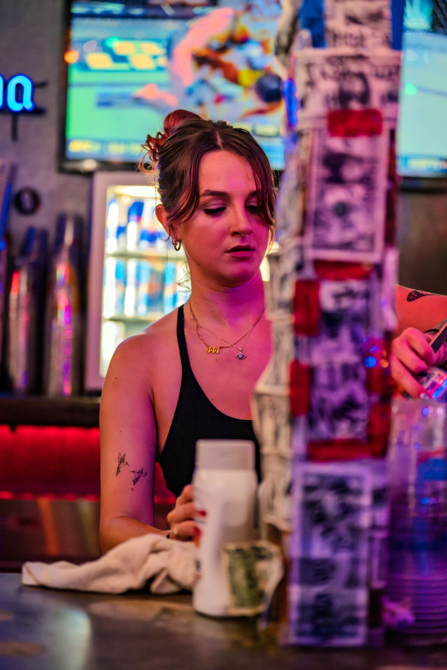 Female bartender in black tank top wiping bar counter under neon lights in a lively bar setting.