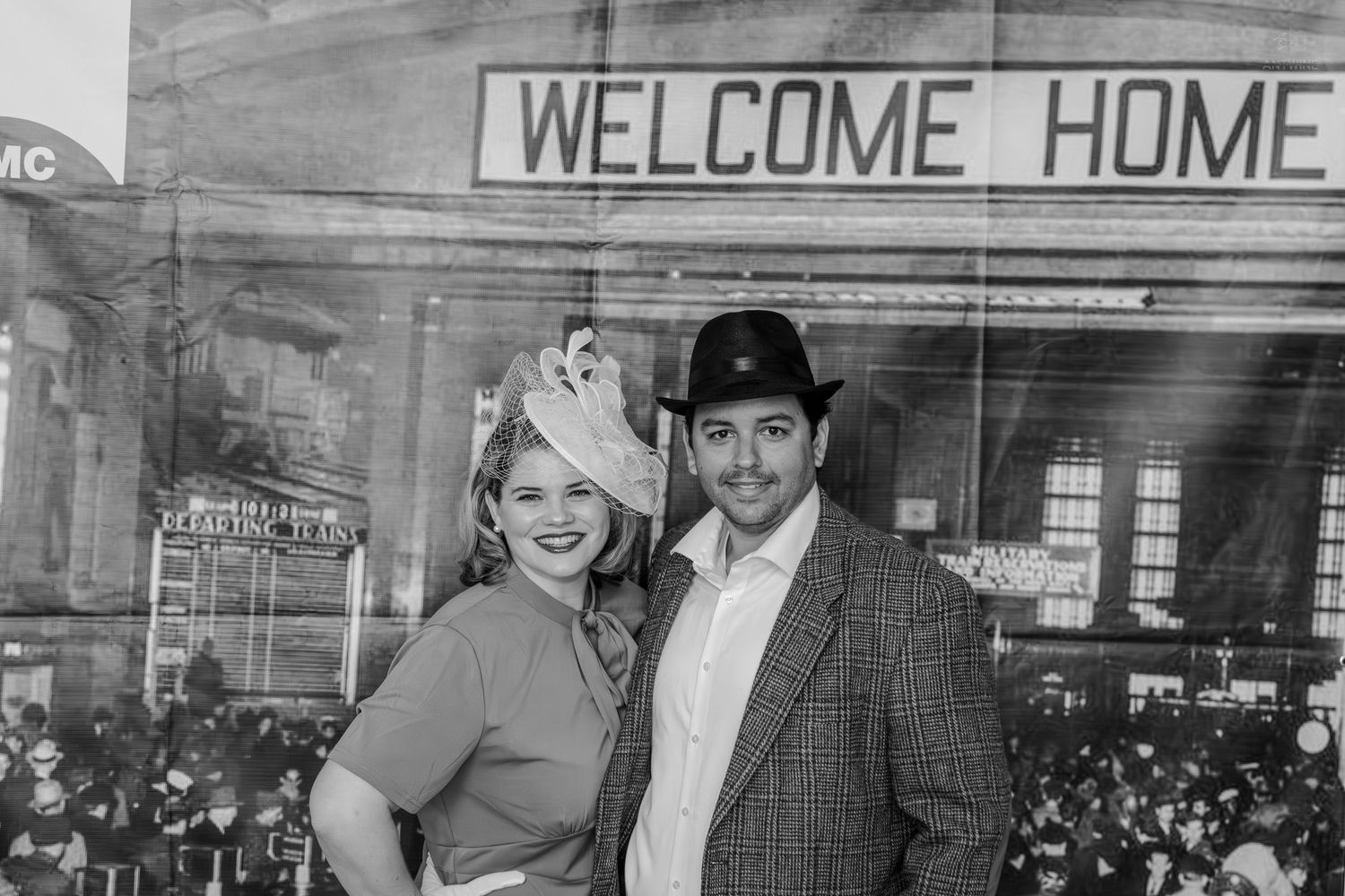 Black and white photo of a couple in vintage attire posing in front of a Welcome Home sign at a train station.