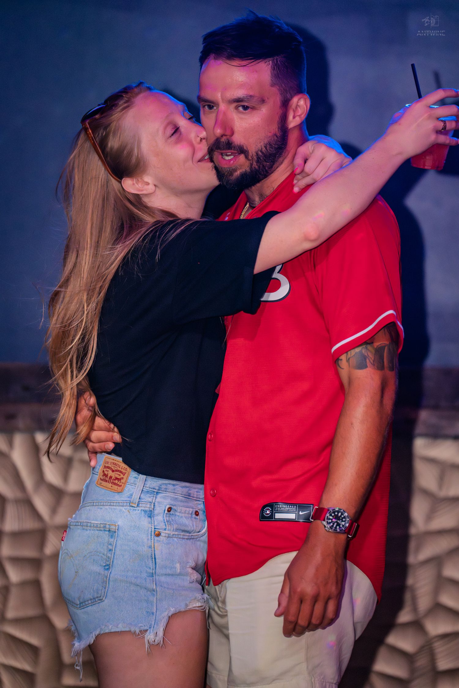 Couple embracing and taking a selfie at a nightclub, woman in black top and denim shorts, man in red jersey.