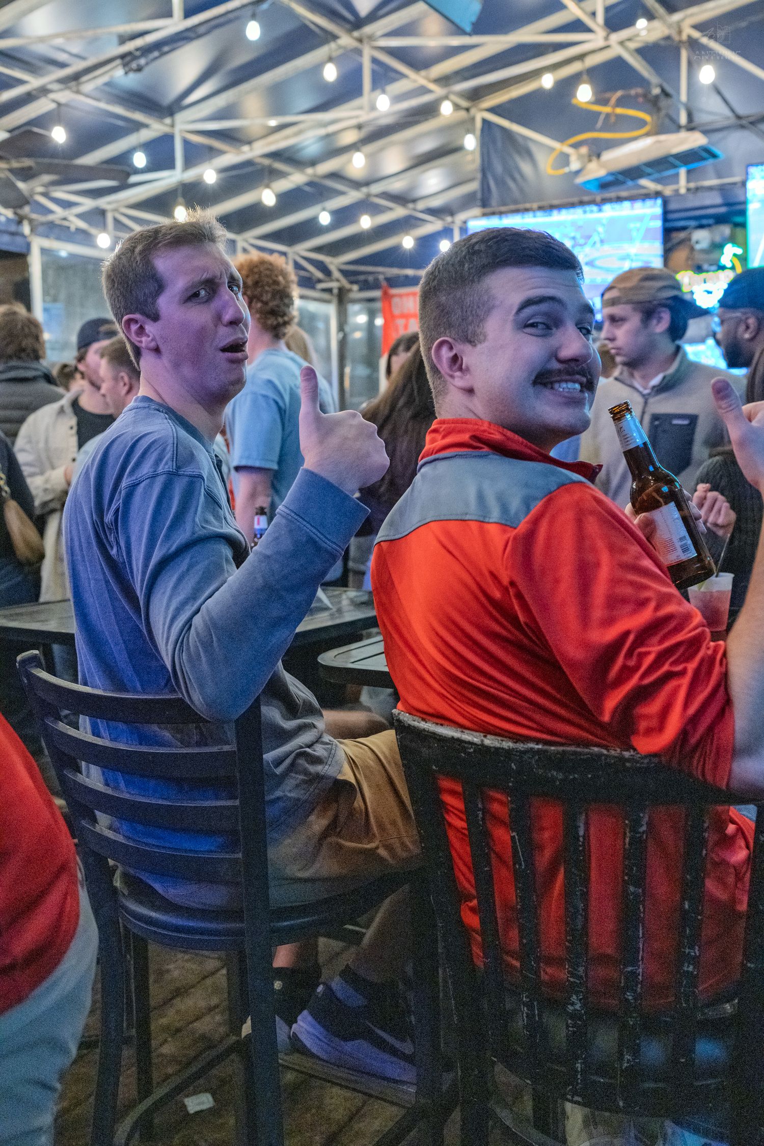 Two young men at a bar, one giving thumbs up and smiling at the camera holding a beer bottle.