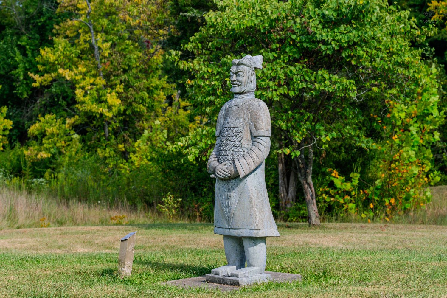 Ancient Chinese terracotta warrior statue replica standing on grass with lush green trees in the background.