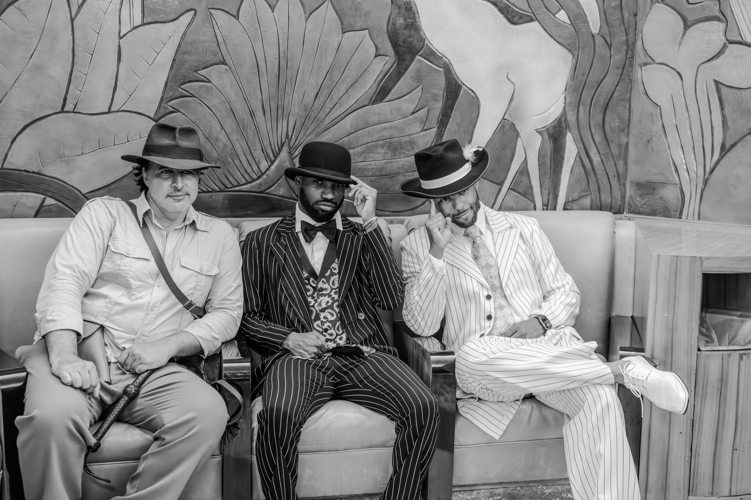 Three stylishly dressed men in vintage suits and hats sitting on a bench in front of an art deco mural.