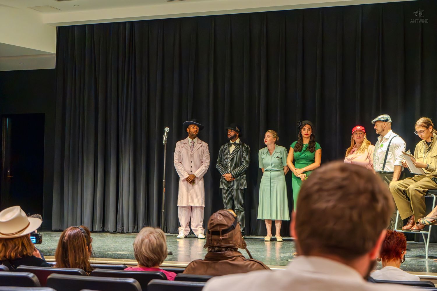 Theater cast members in period costumes take a bow on stage during a dramatic performance before a live audience.