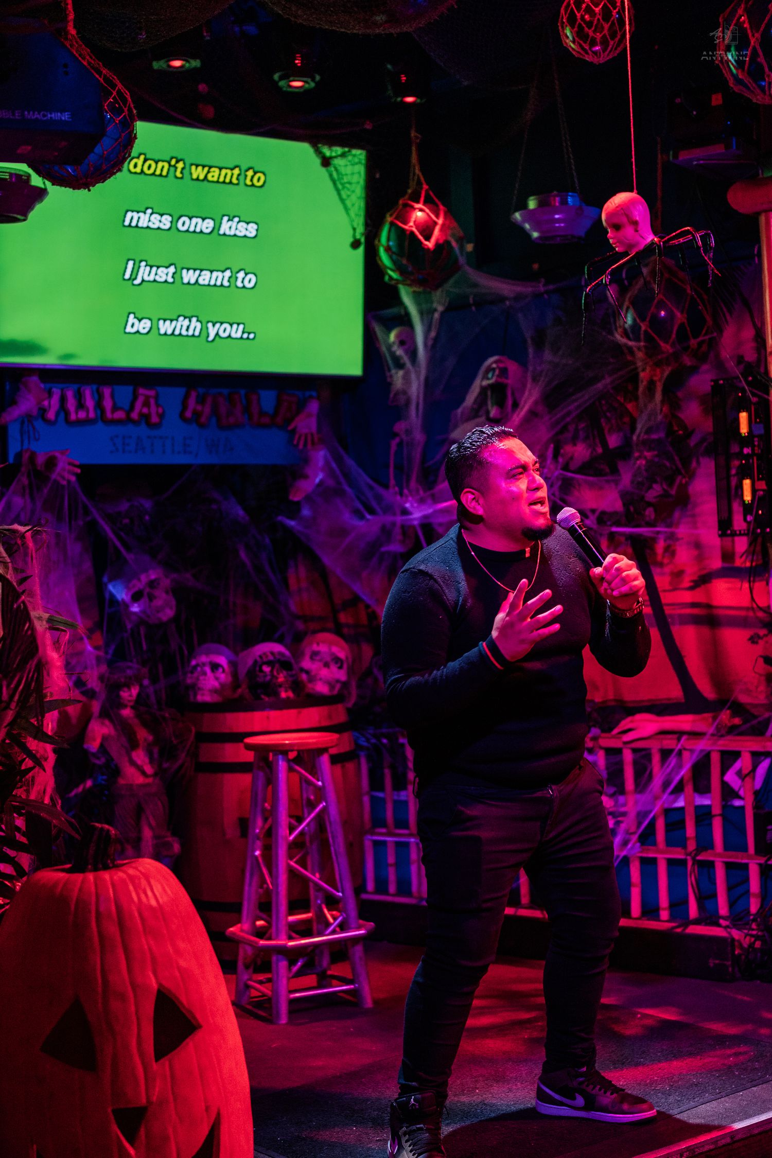 Man singing karaoke on stage at Halloween-themed bar with green lyric screen and skull decorations.