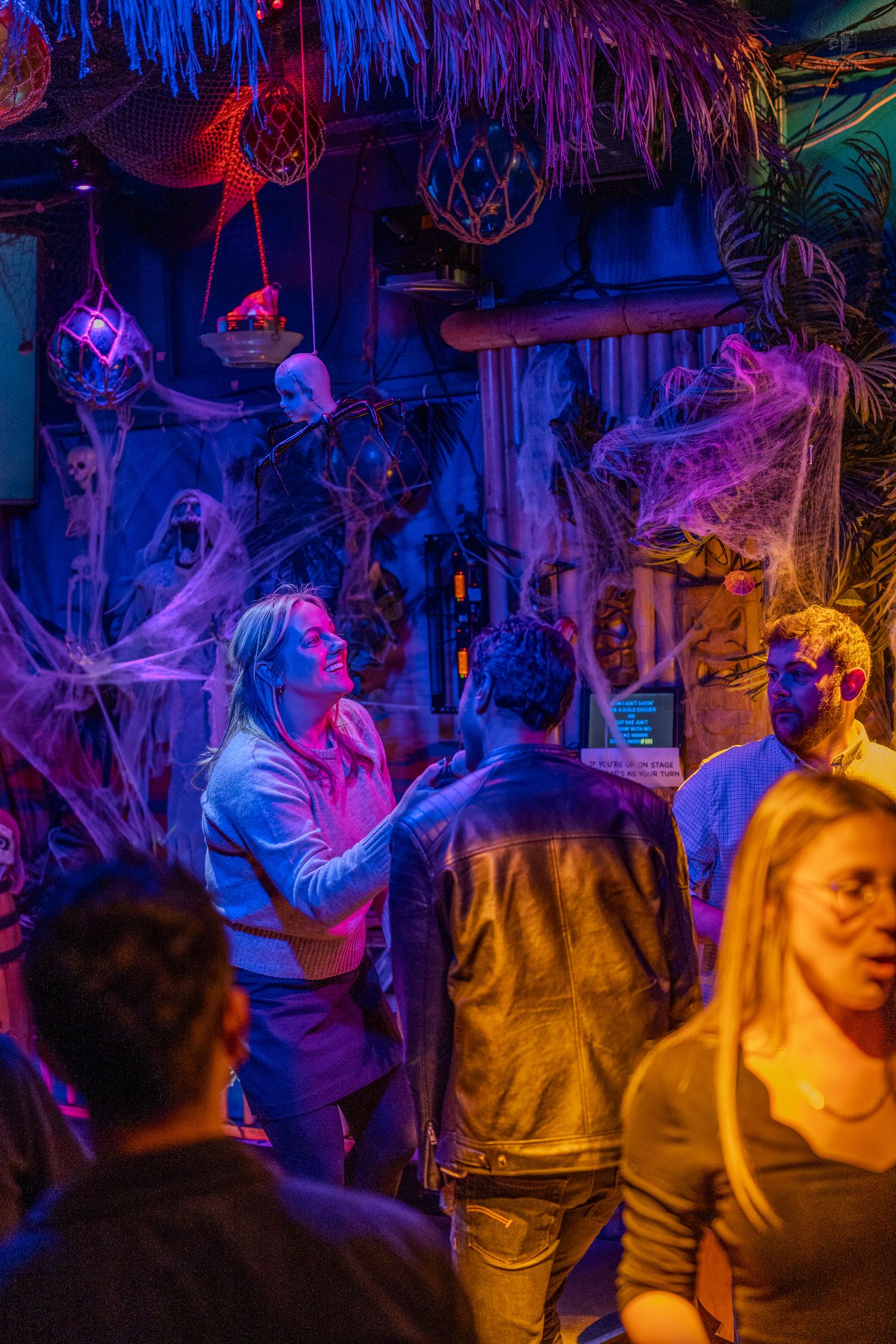 People explore a spooky Halloween-themed bar with cobwebs, skulls, and colorful purple and blue neon lighting.