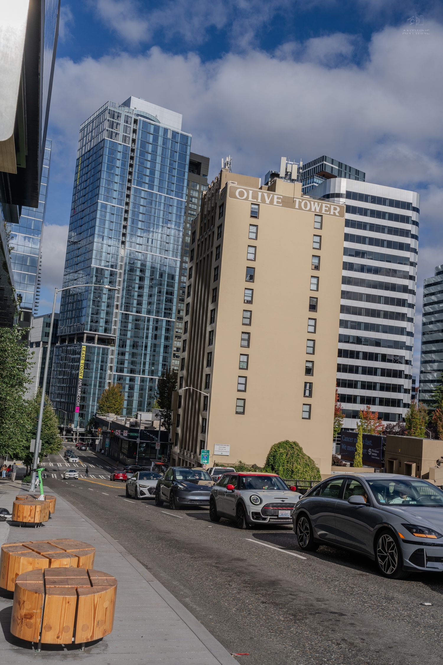 Urban street view of Seattle with Olive Tower building, modern glass skyscrapers, and parked cars under cloudy sky.