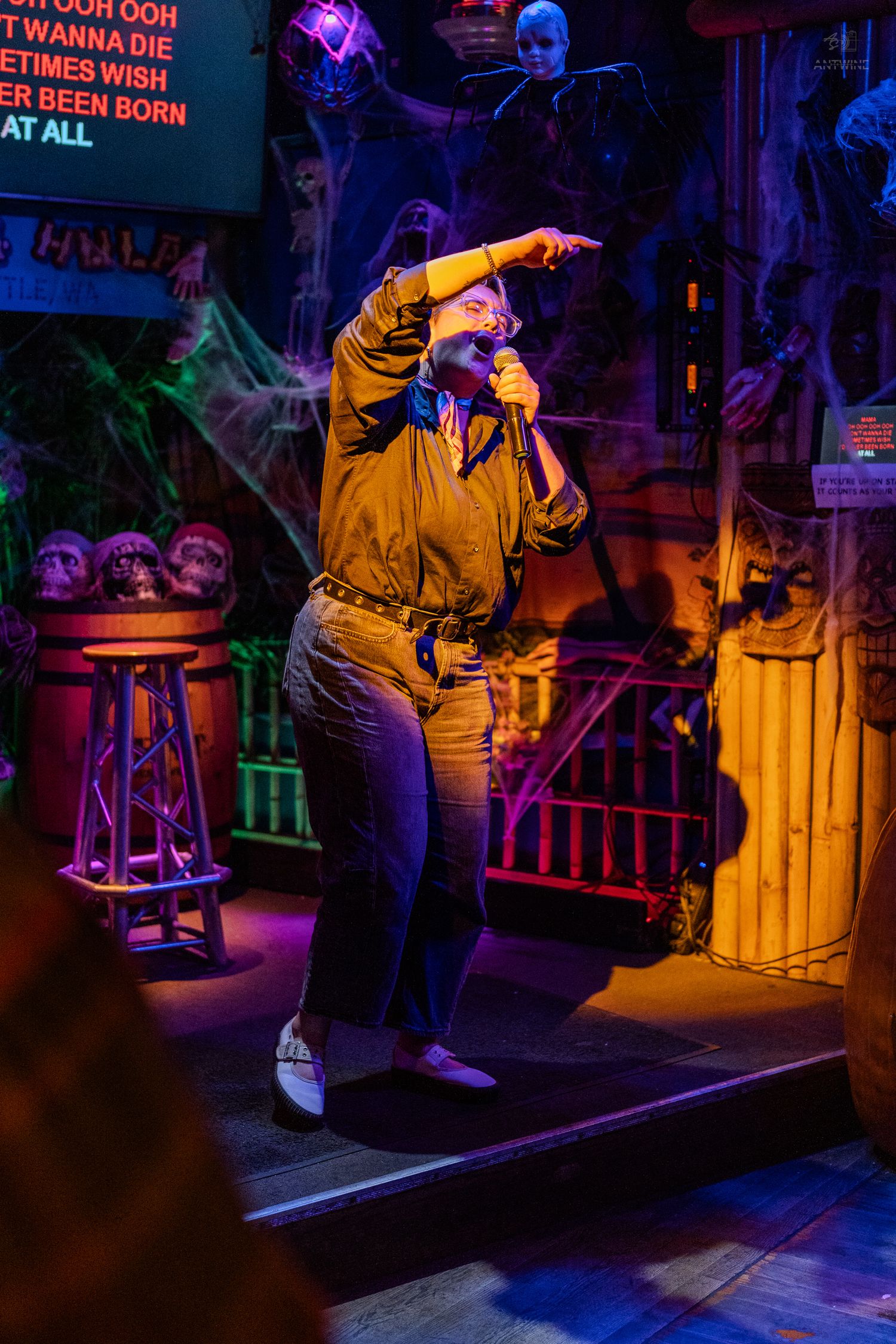 Person singing karaoke on Halloween-decorated stage with colorful lighting, cobwebs, and skulls in background.