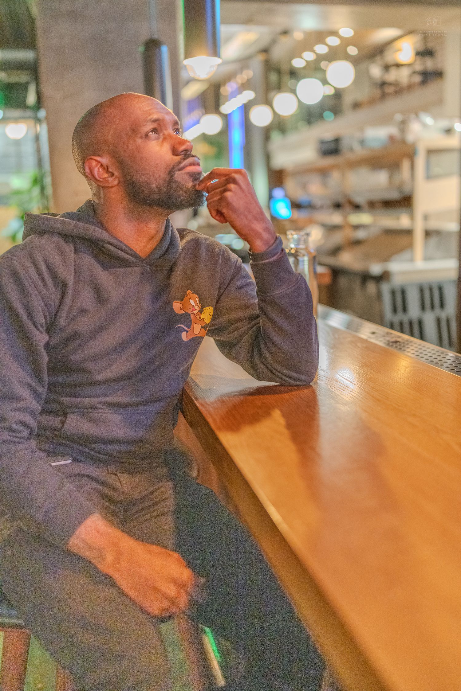 A thoughtful bald Black man with a beard sits at a wooden bar counter in a modern restaurant, resting his chin on his hand.