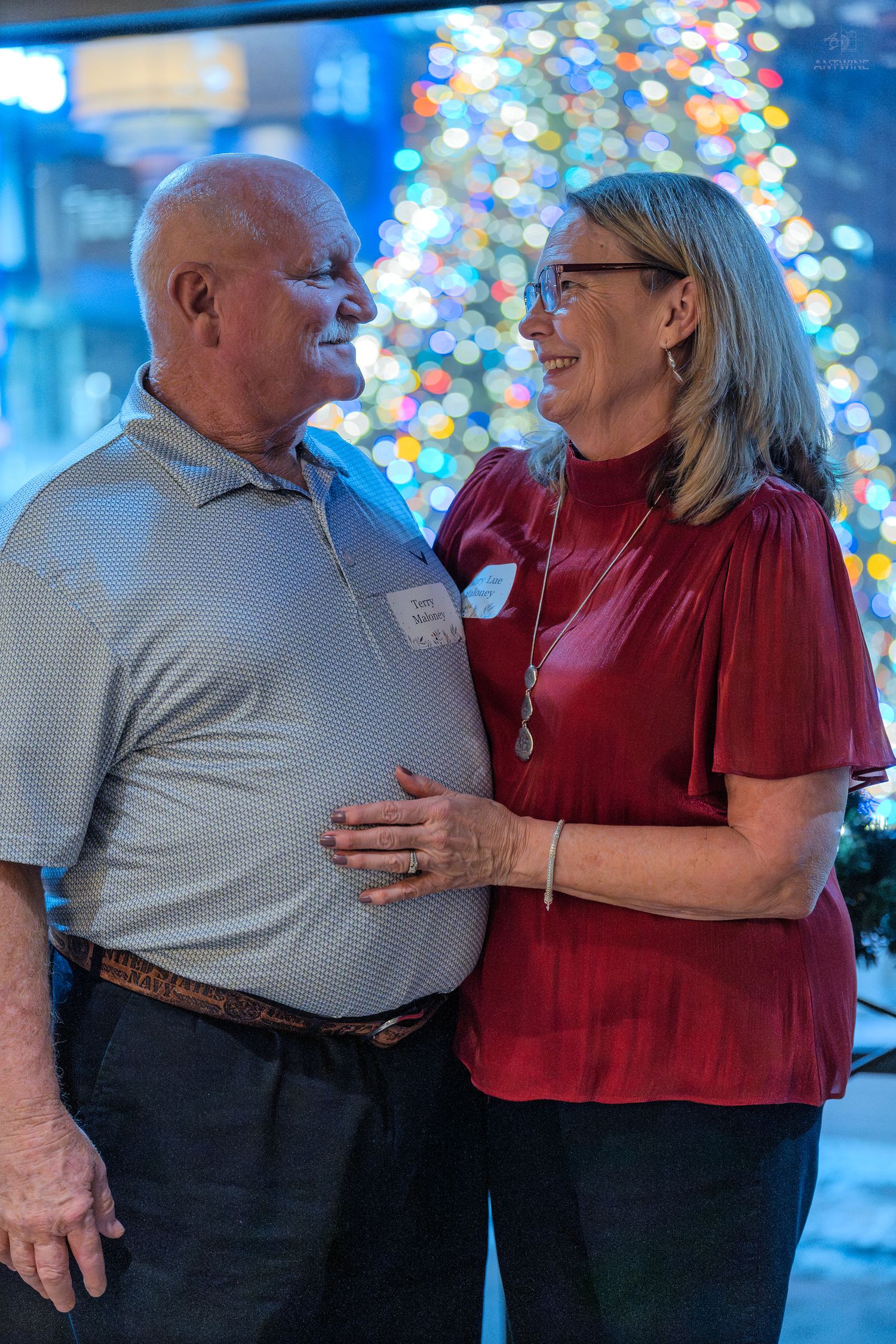 Couple smiling at each other at a festive holiday party with a decorated Christmas tree in the background.