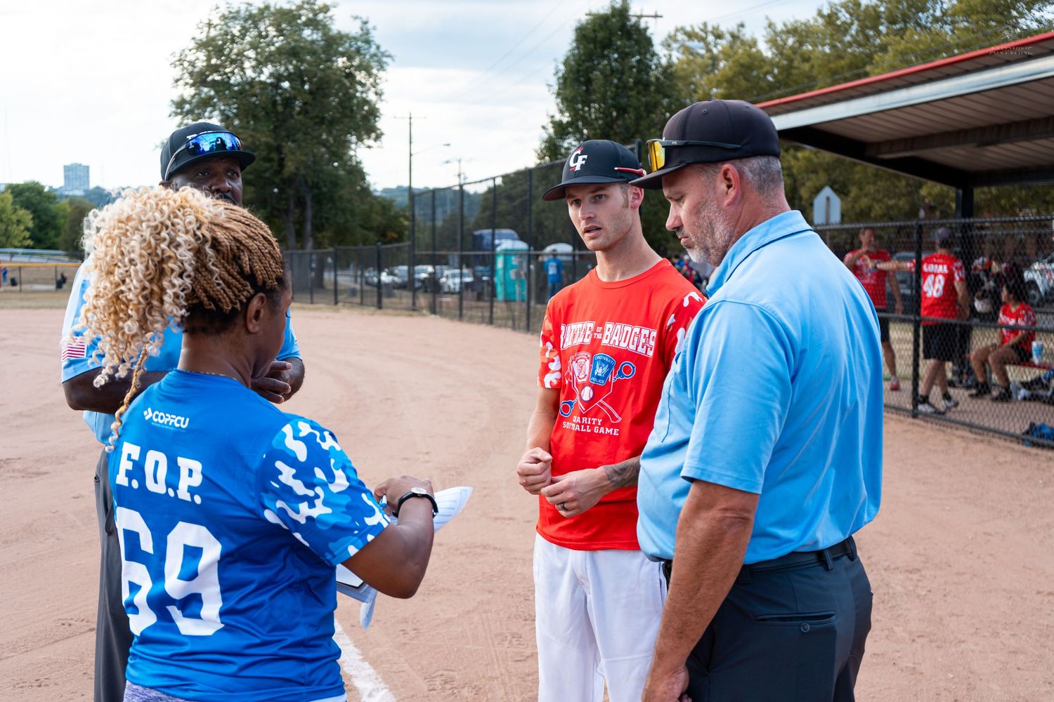 A group of people including a player in a blue FOP jersey discuss at a baseball field during a game.