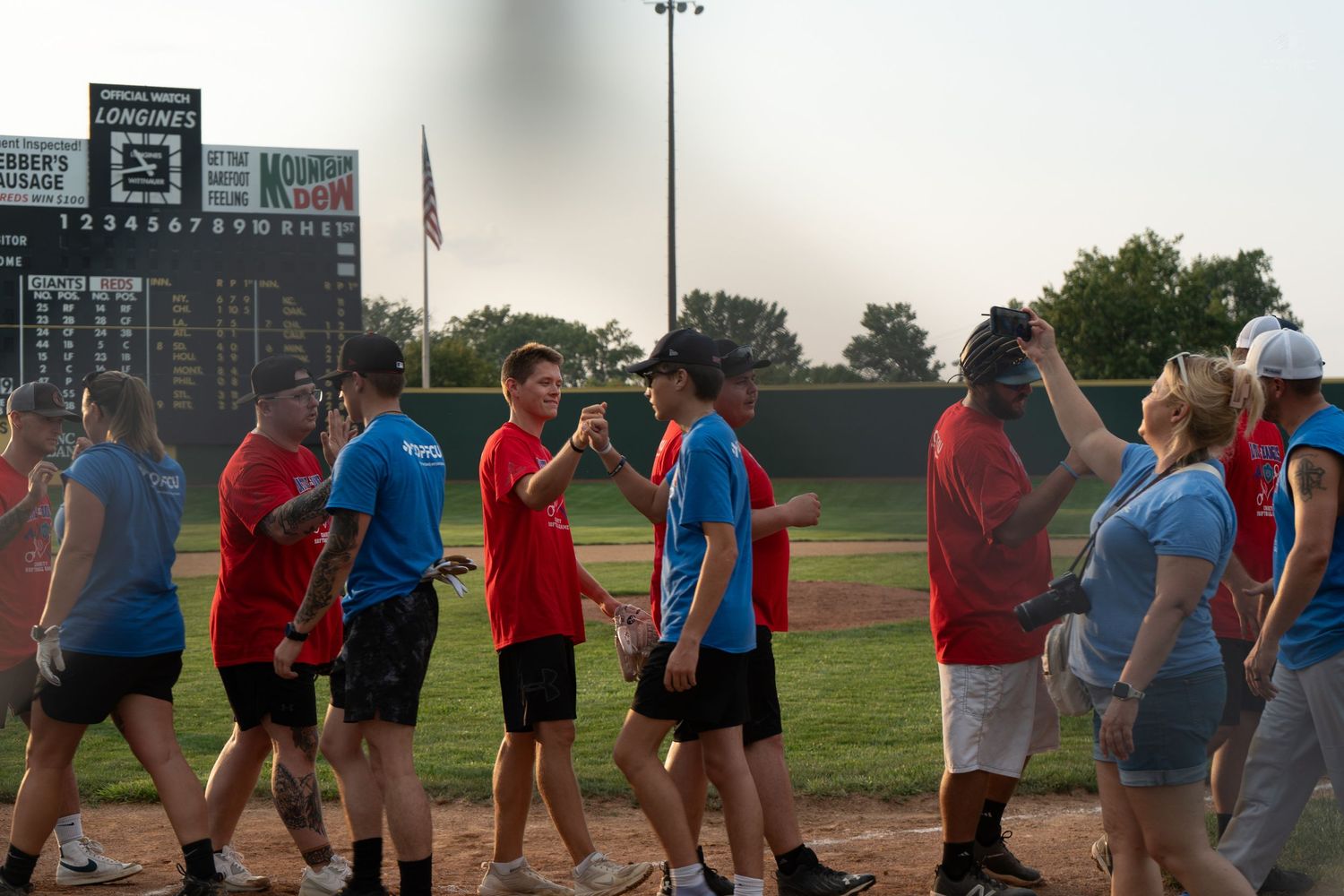 Teams shake hands on a baseball field after a recreational softball game under evening skies.