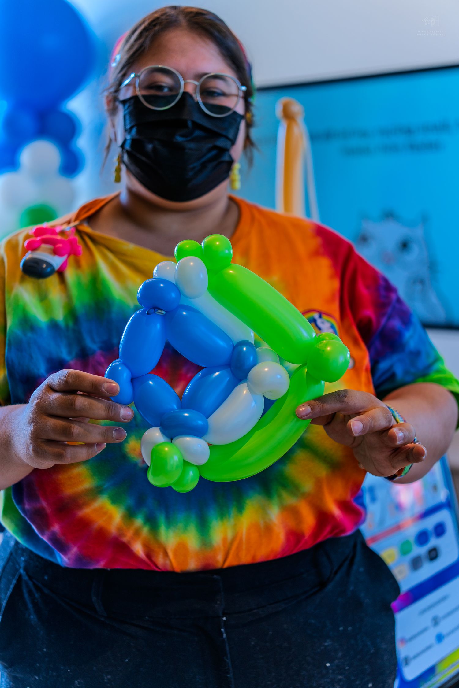 Woman in tie-dye shirt and black mask holds a creative balloon sculpture in blue and green colors.