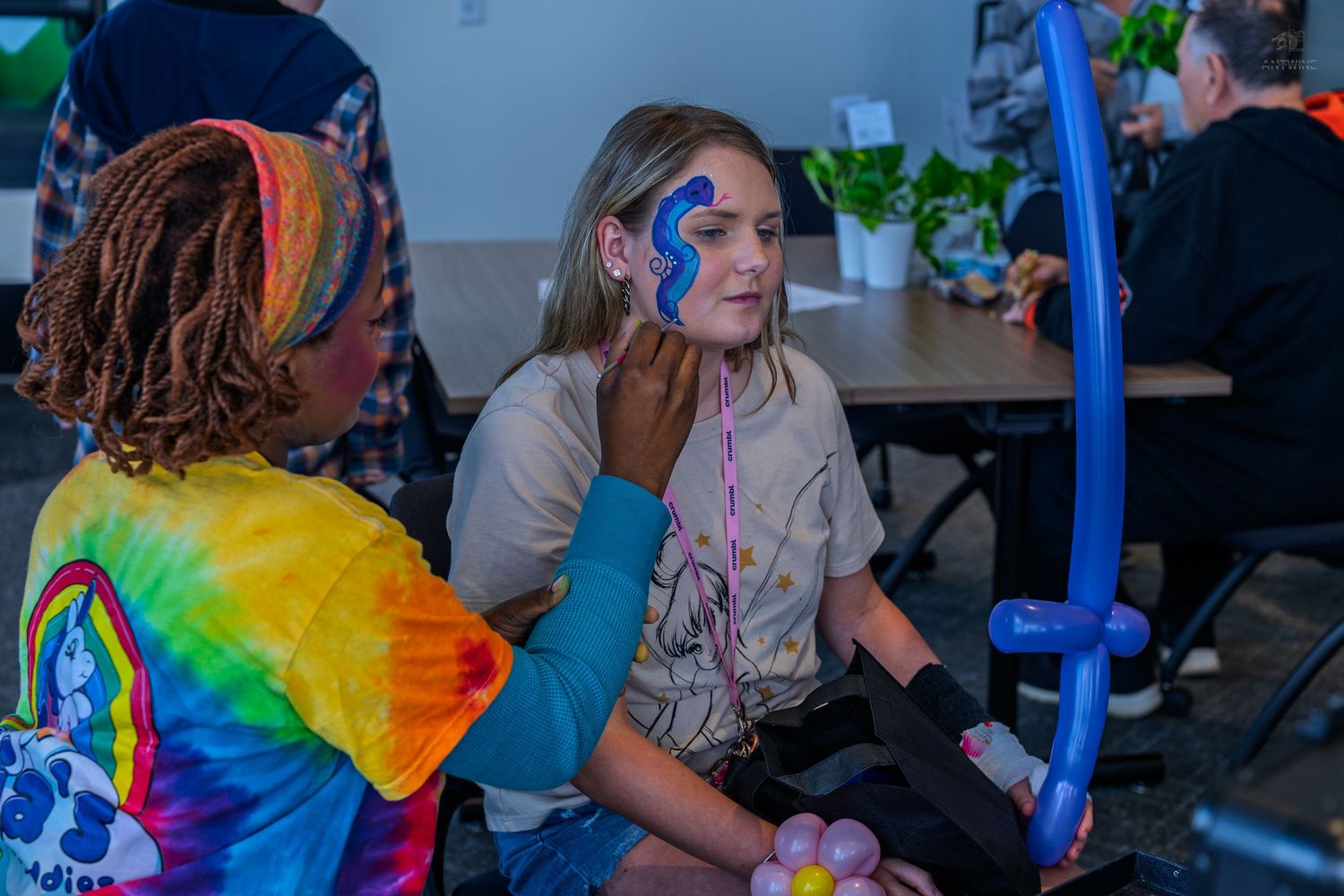 A face painter in tie-dye applies blue butterfly face paint to a young girl at a festive event.