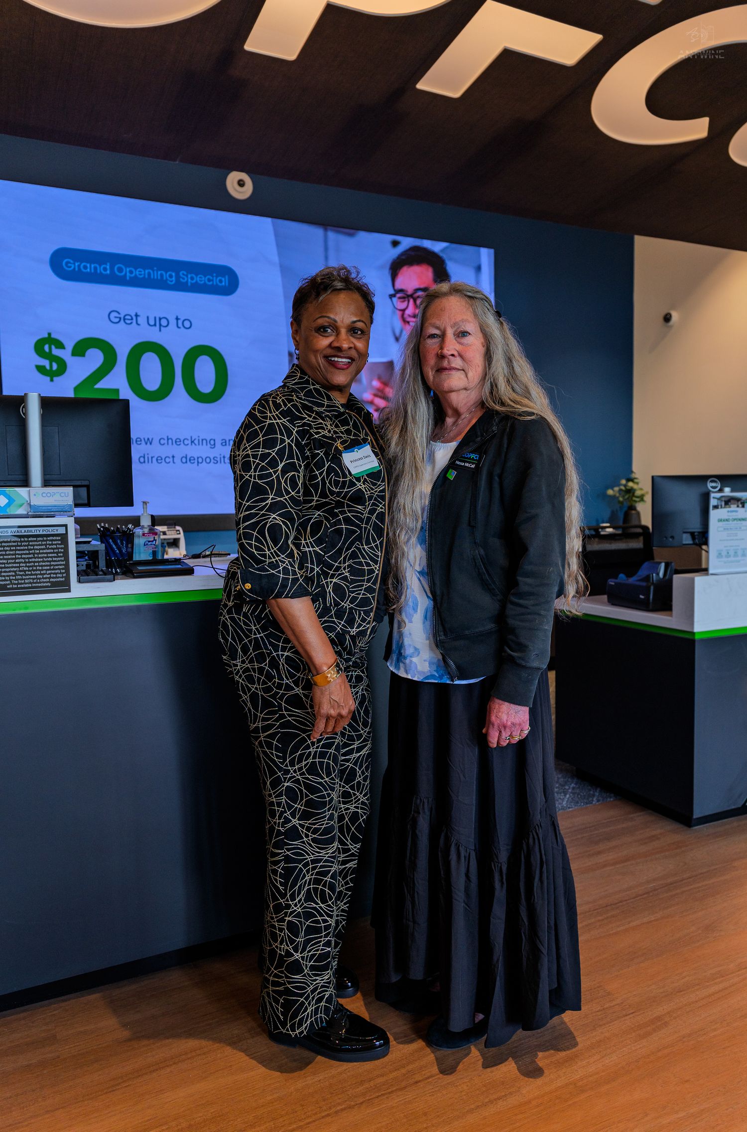 Two women pose together at COPFCU bank grand opening with $200 new checking account promotion displayed behind them.