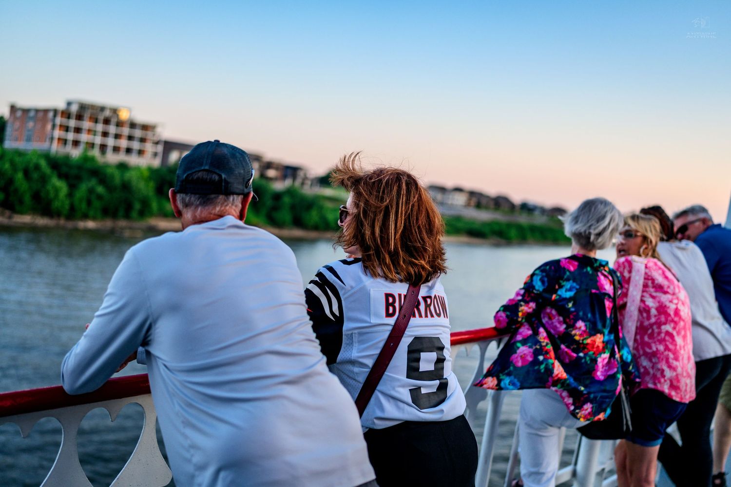 Group of people standing at ship railing watching scenic river sunset view.