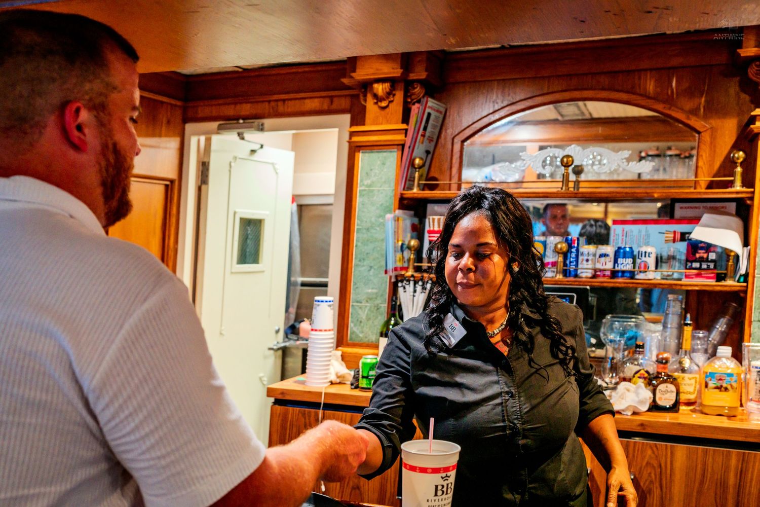 A woman in a black uniform smiles while handing a drink to a man at an indoor bar counter.