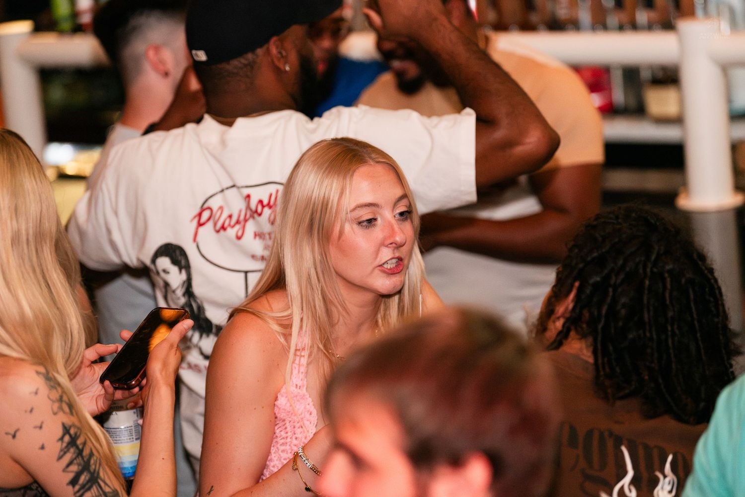 Blonde woman in pink top socializing at a lively bar with friends.