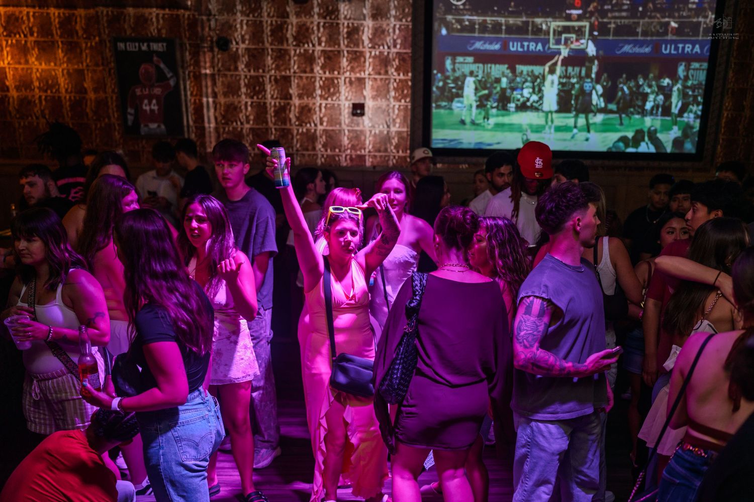 Excited crowd dancing and raising hands in a purple-lit nightclub with a basketball game on screen.
