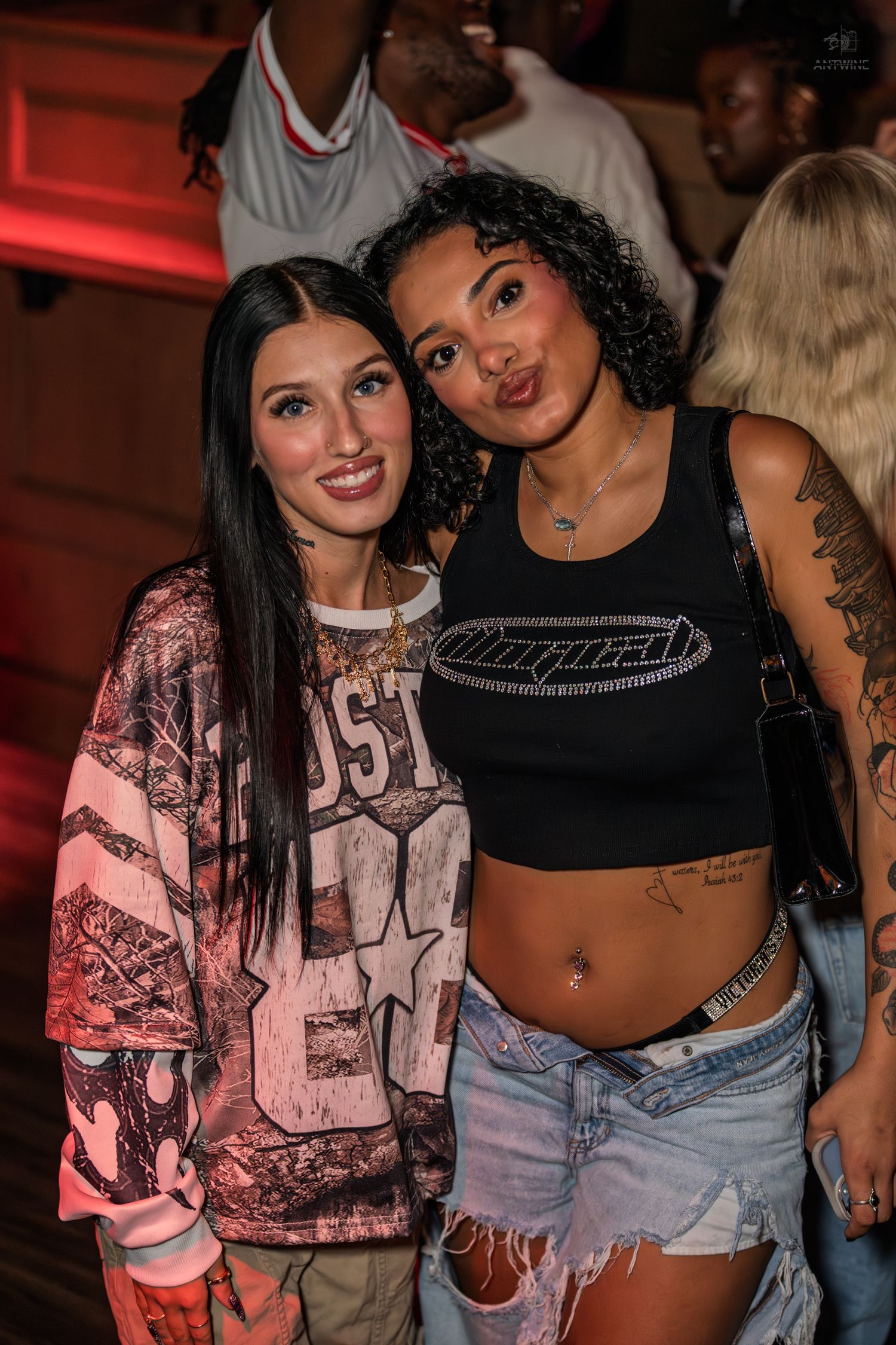 Two women pose together smiling at a nightclub, one in a graphic sweatshirt and the other in a black crop top with tattoos.