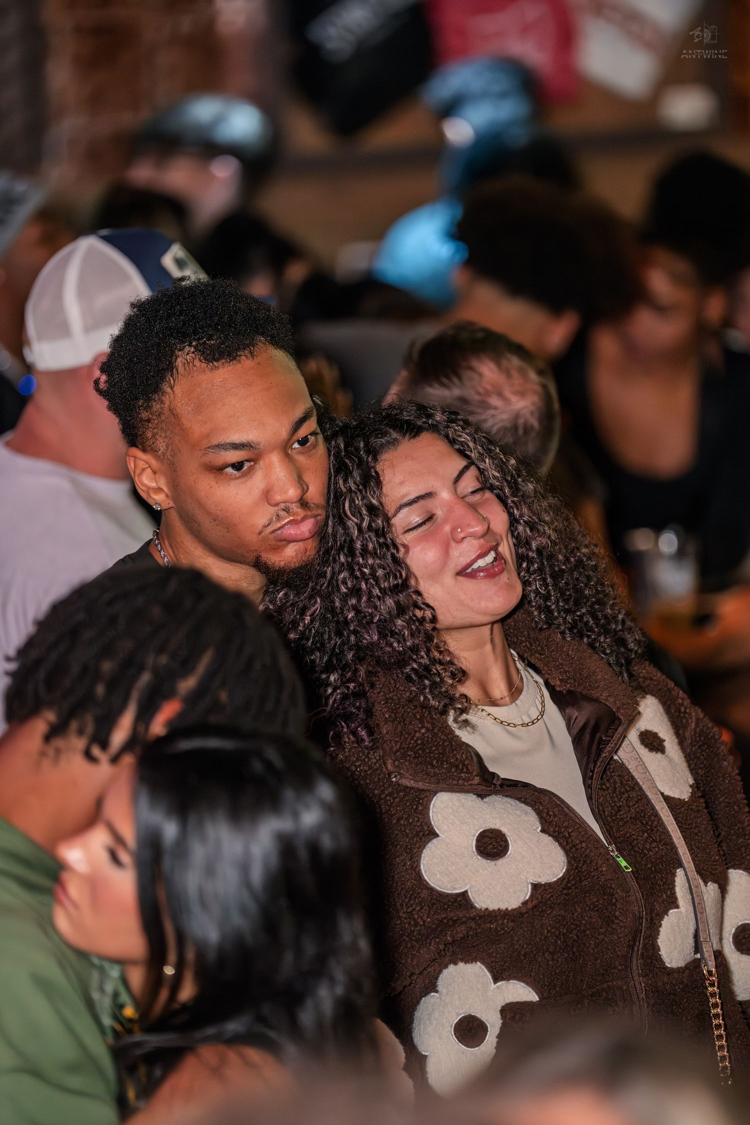 Couple sharing a tender moment at a crowded bar, woman in brown floral jacket laughing joyfully.