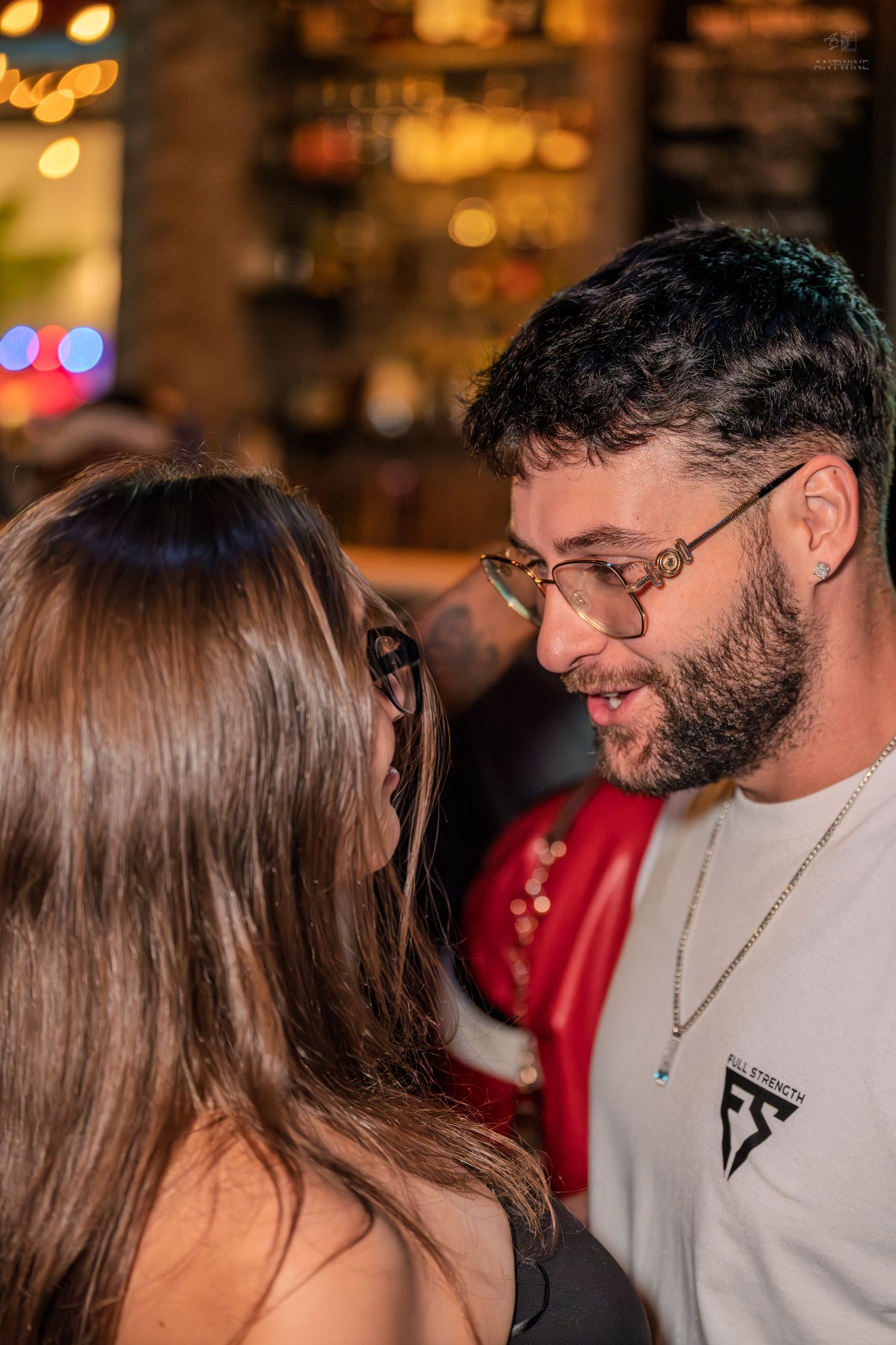 Young bearded man with gold glasses talking closely with a woman at a bar.