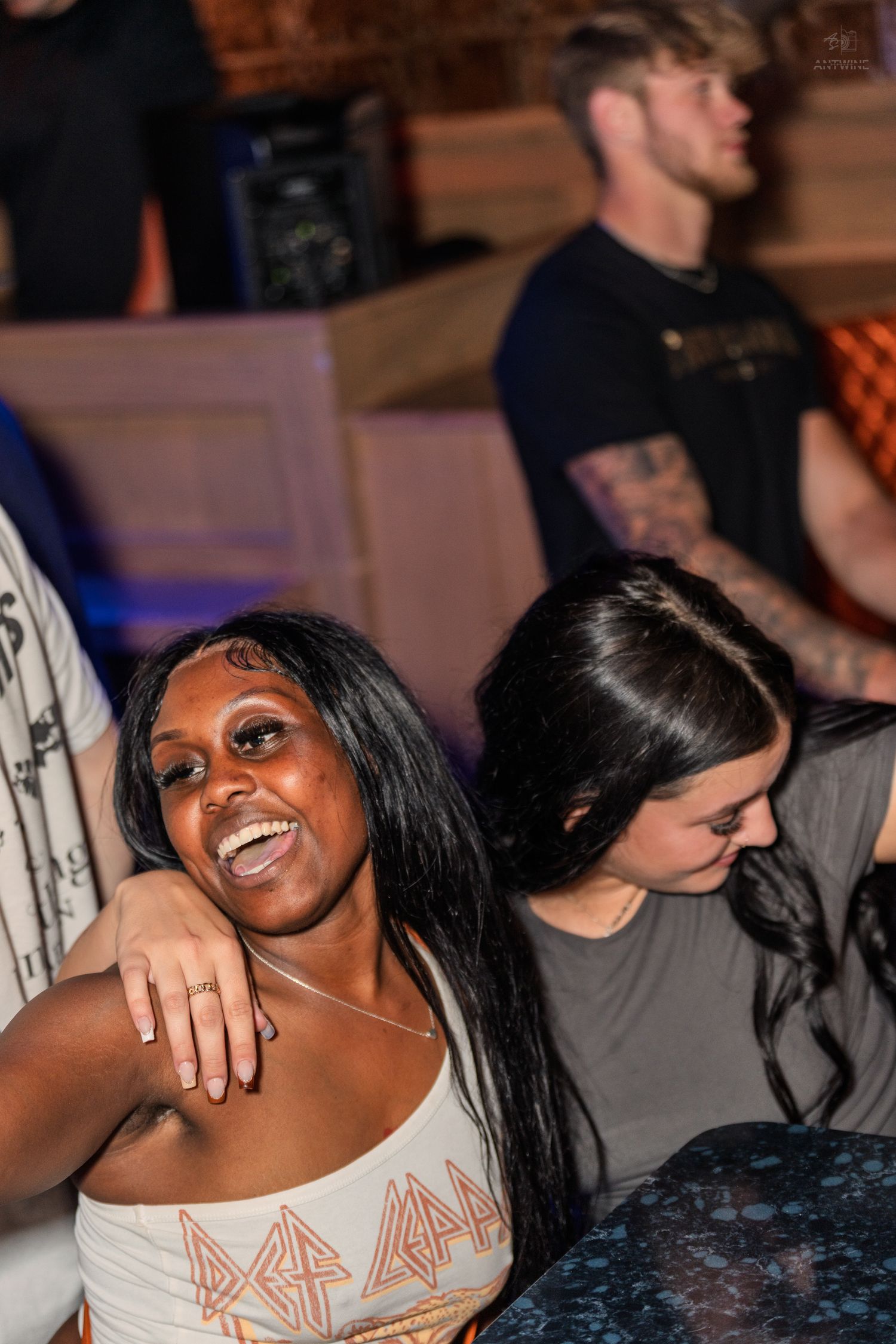 Smiling woman in Def Leppard top laughing joyfully with friend at an indoor nightclub event.
