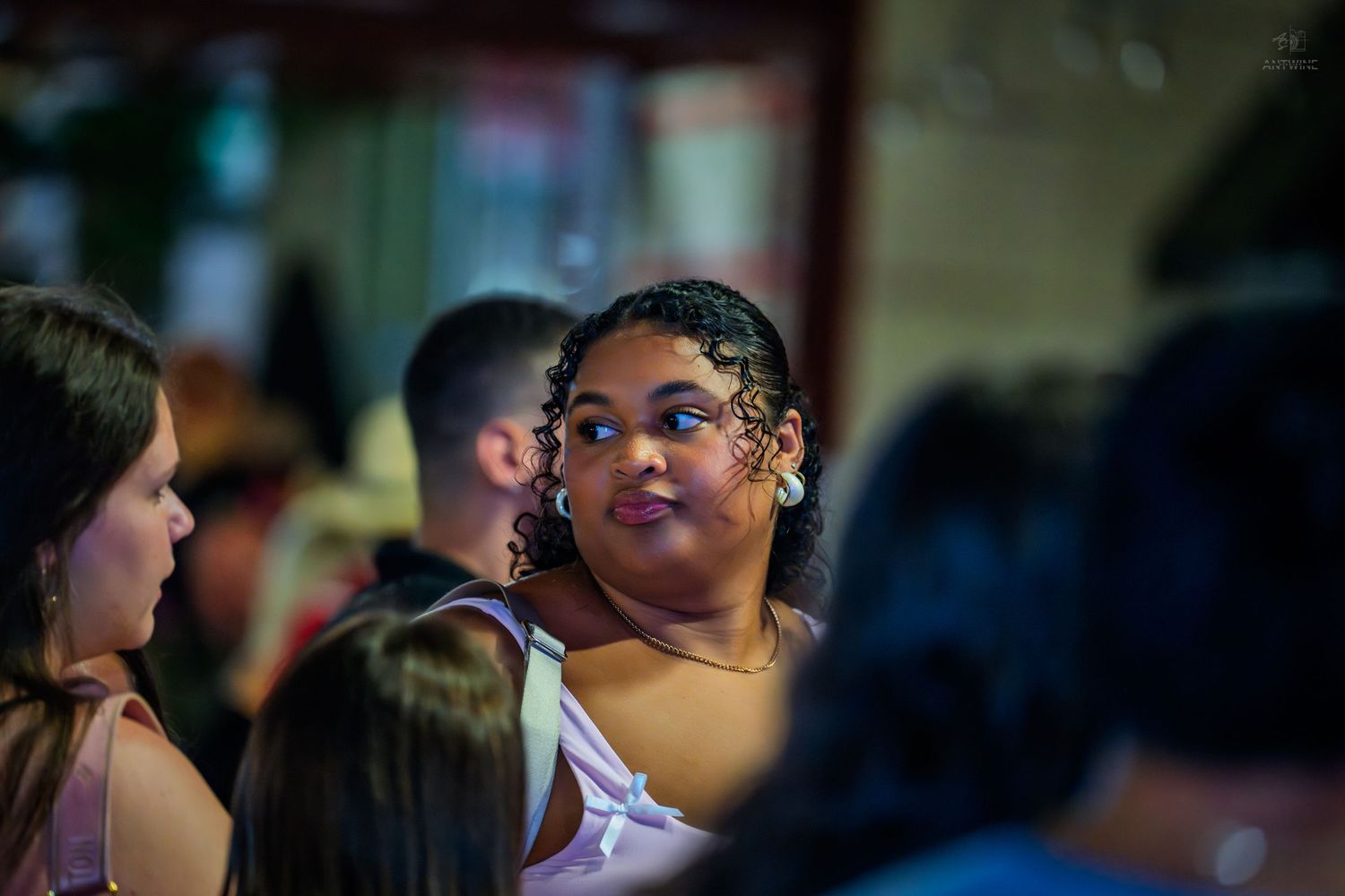 Young woman with curly hair looking sideways in a crowded indoor venue.