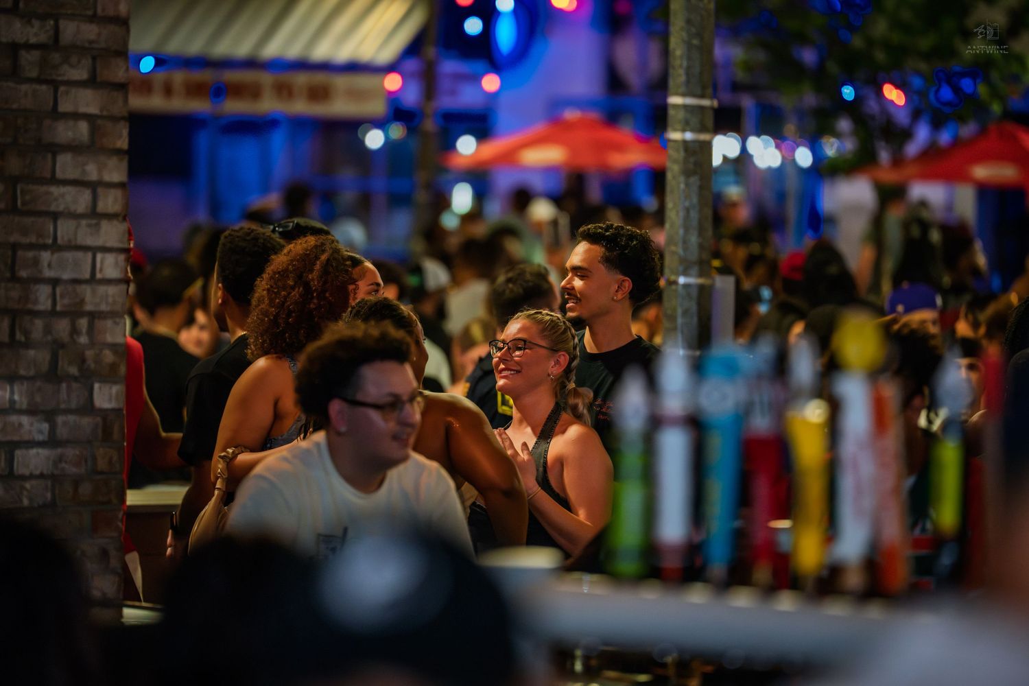 Crowded outdoor bar patio at night with colorful neon lights and groups of people socializing.