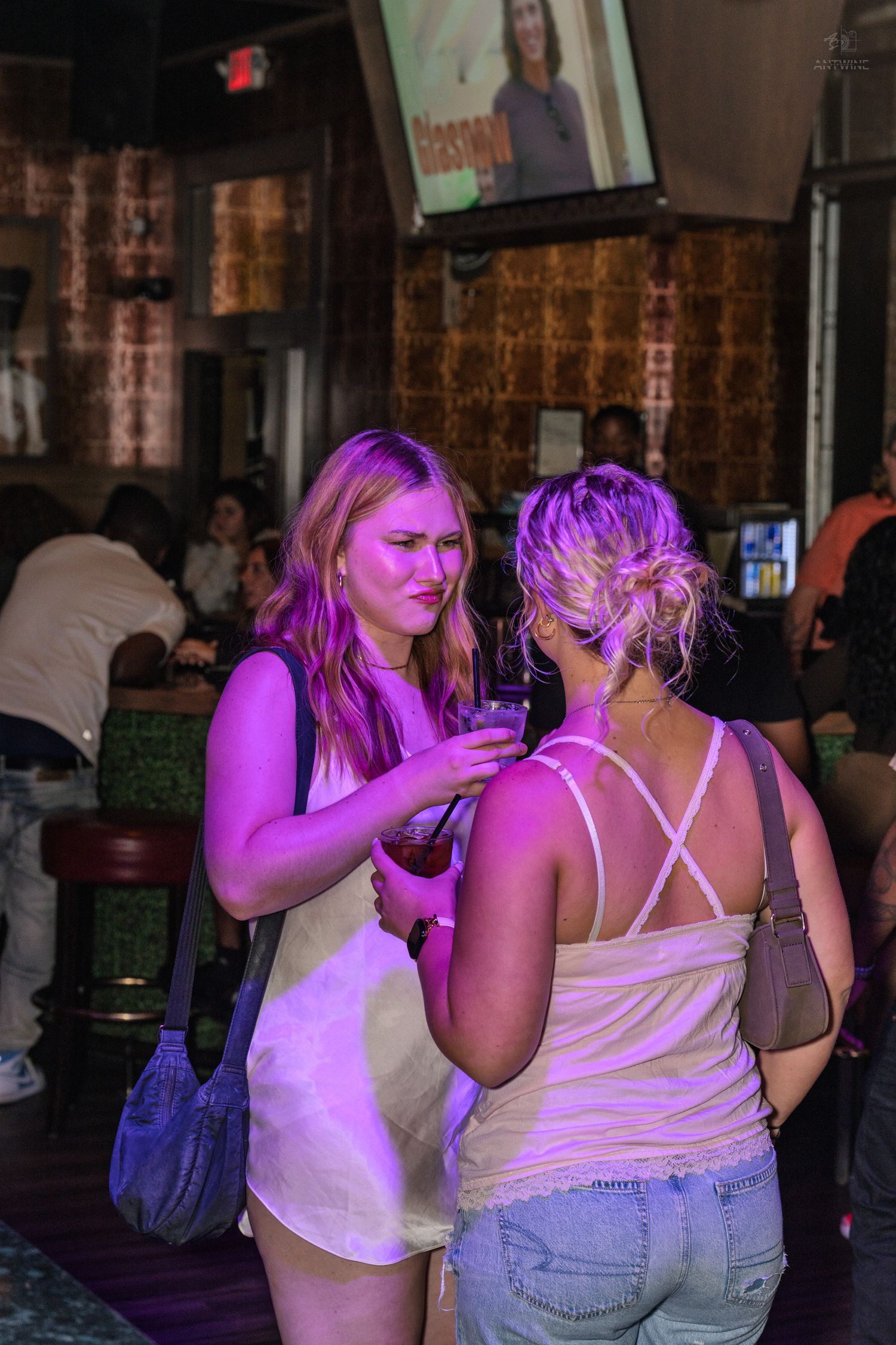 Two women talking under purple neon lights at a crowded bar, one in white dress, one in pink top.