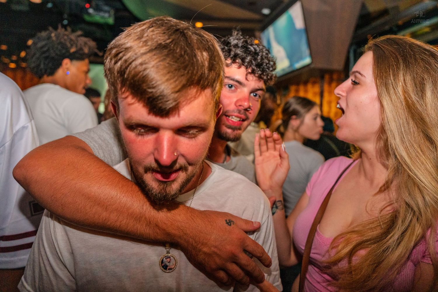 Three young adults laughing and hugging at a busy bar with warm lighting.