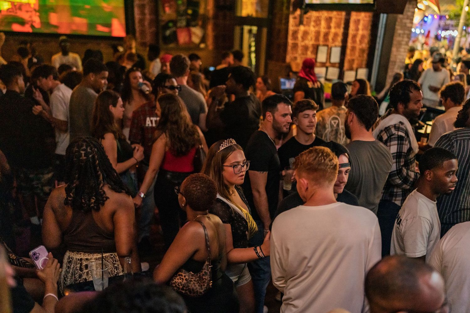 Large crowd of young adults socializing at an outdoor bar venue with vibrant neon signage.