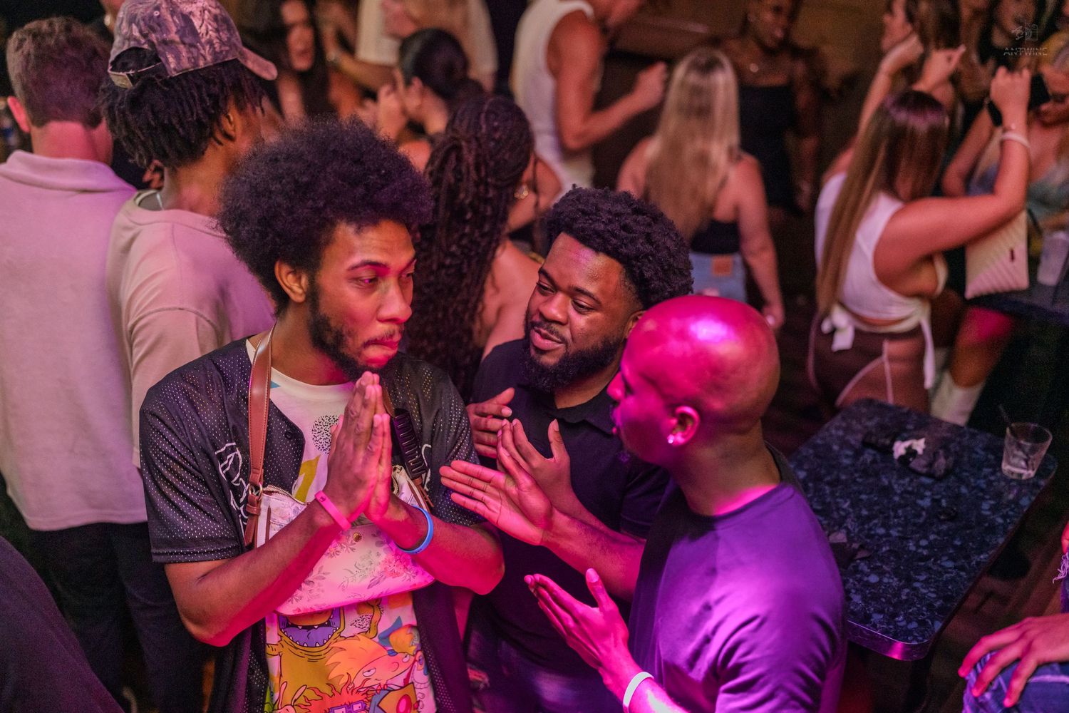 Young men conversing with prayer hand gestures under pink neon lights at a nightclub.