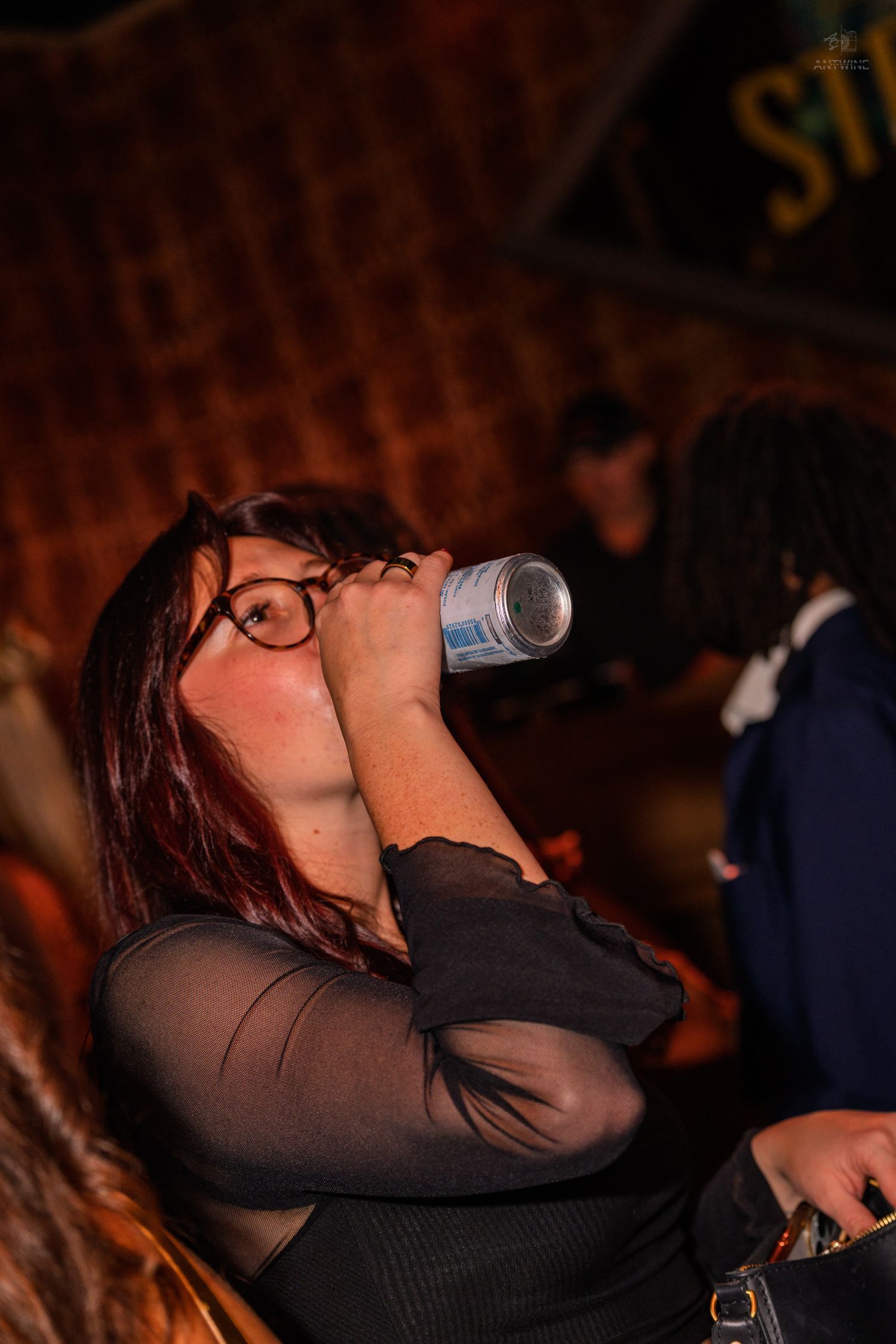 Young woman with glasses drinking from a can at a dimly lit nightclub party.