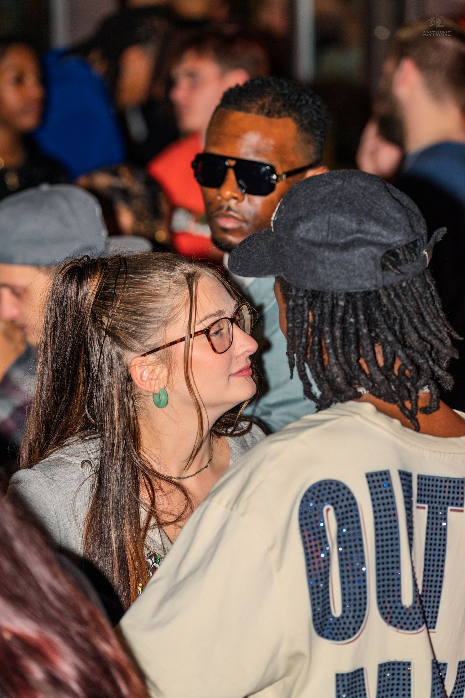 Woman with green earrings and glasses conversing with a man in a sequined jacket at a party.