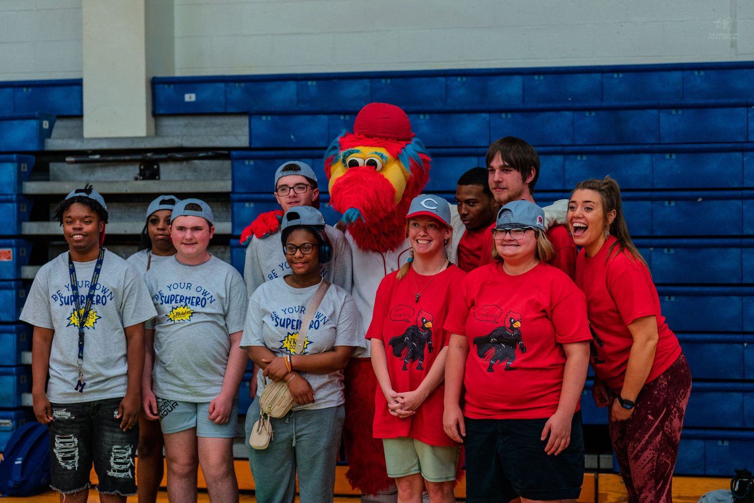 Group of students in red shirts pose with a red bird mascot in a school gymnasium.