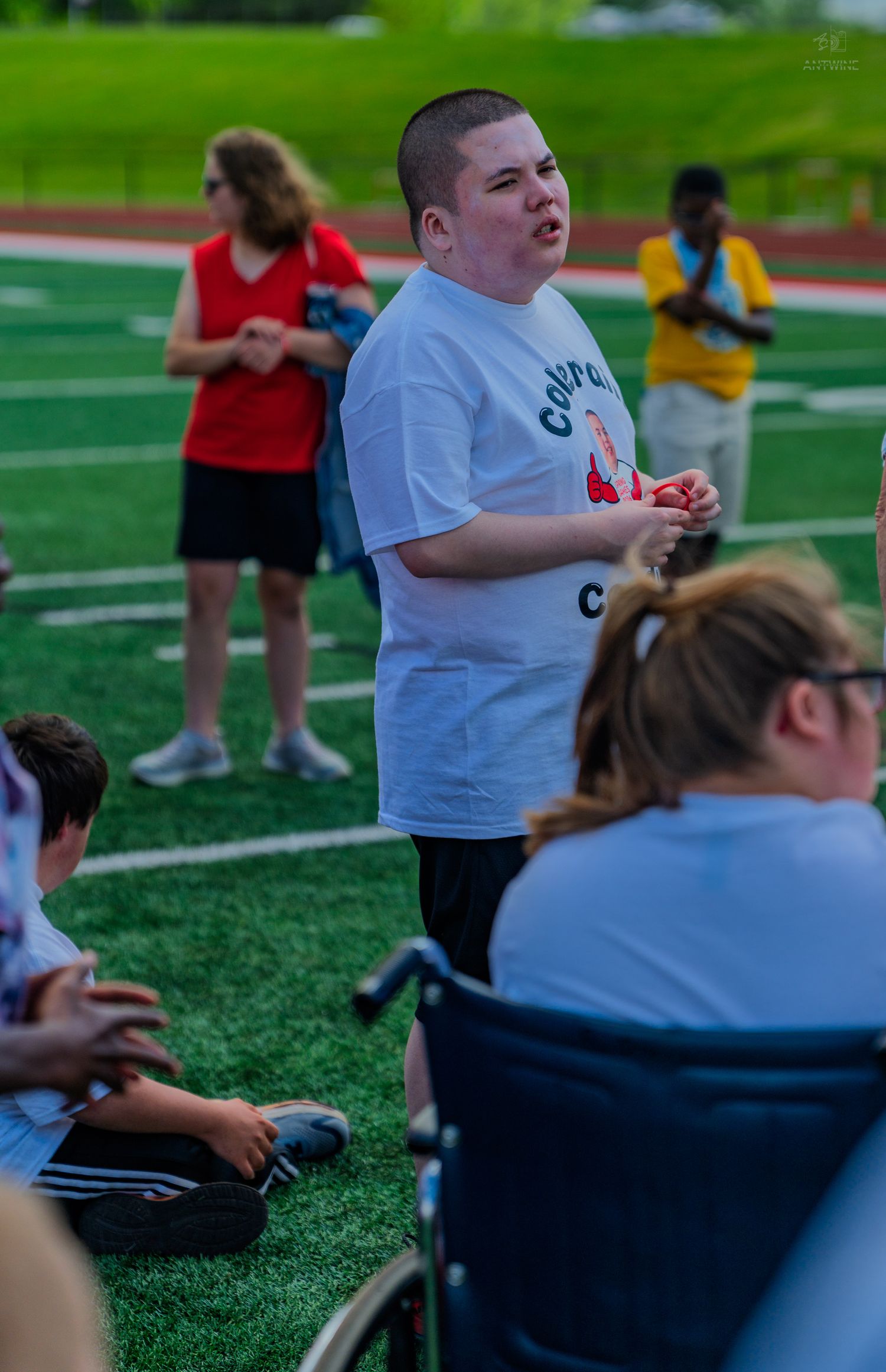 Young person in white t-shirt stands on green turf field surrounded by others, including someone in a wheelchair.