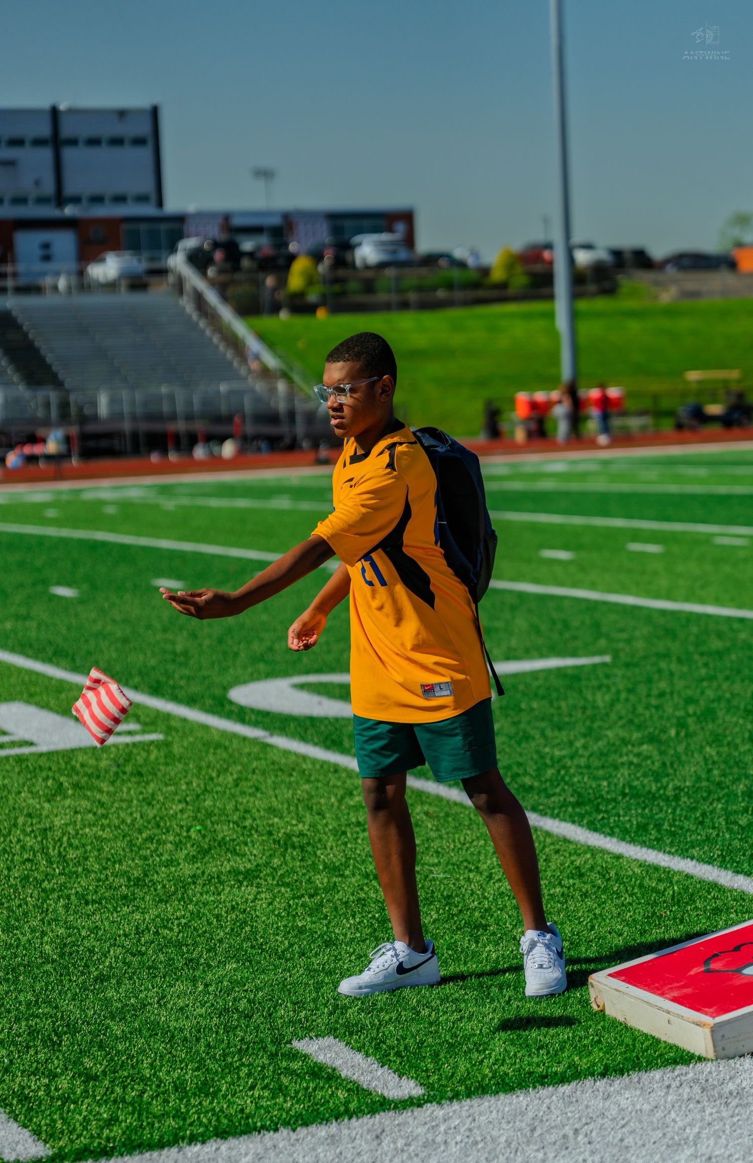Young man in yellow jersey tossing a cornhole bag on a green turf football field during an outdoor game.