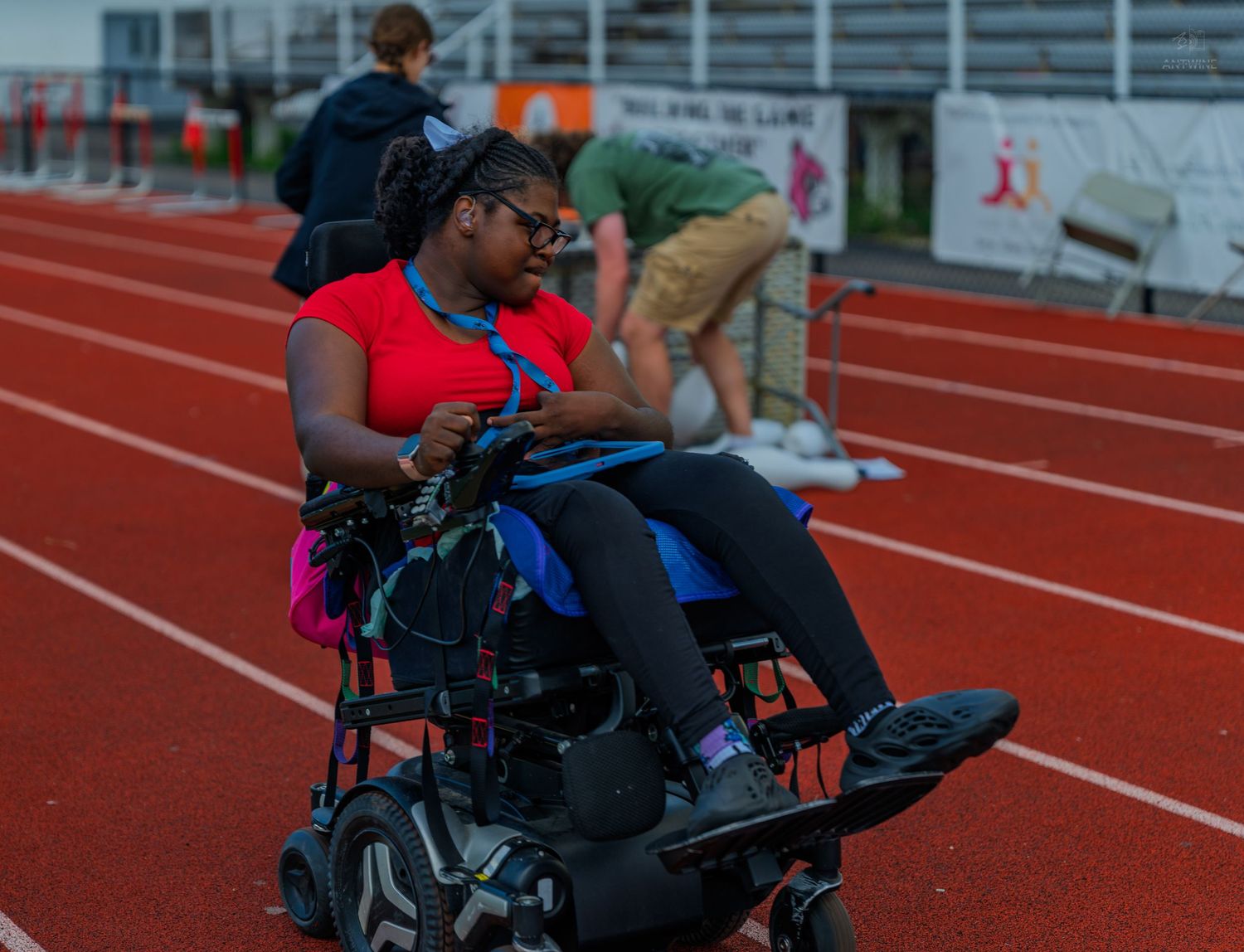 Woman in red shirt using a power wheelchair on a red running track at an outdoor athletics stadium.