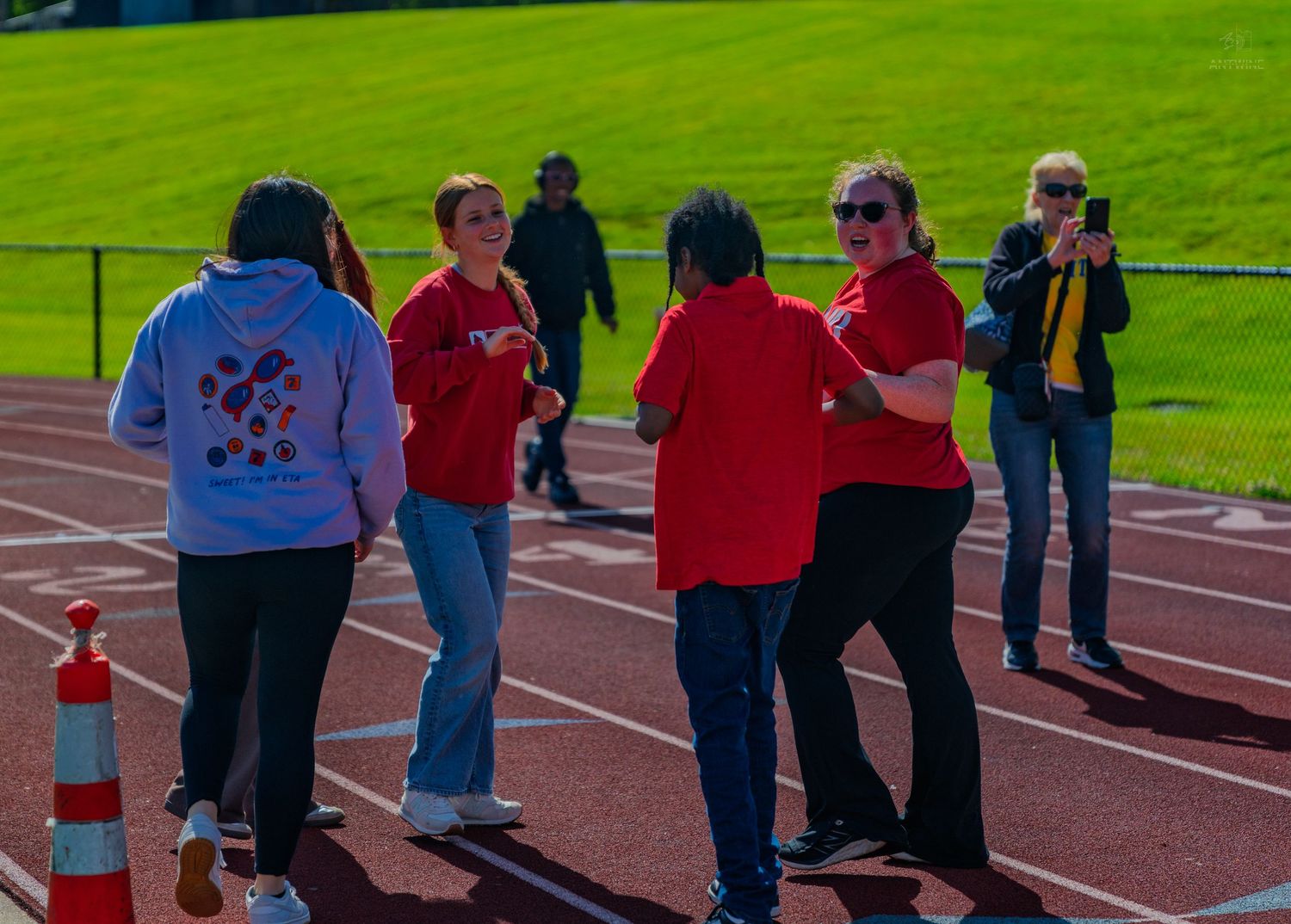 Students in red shirts interact on a running track during an outdoor special needs athletic event on a sunny day.