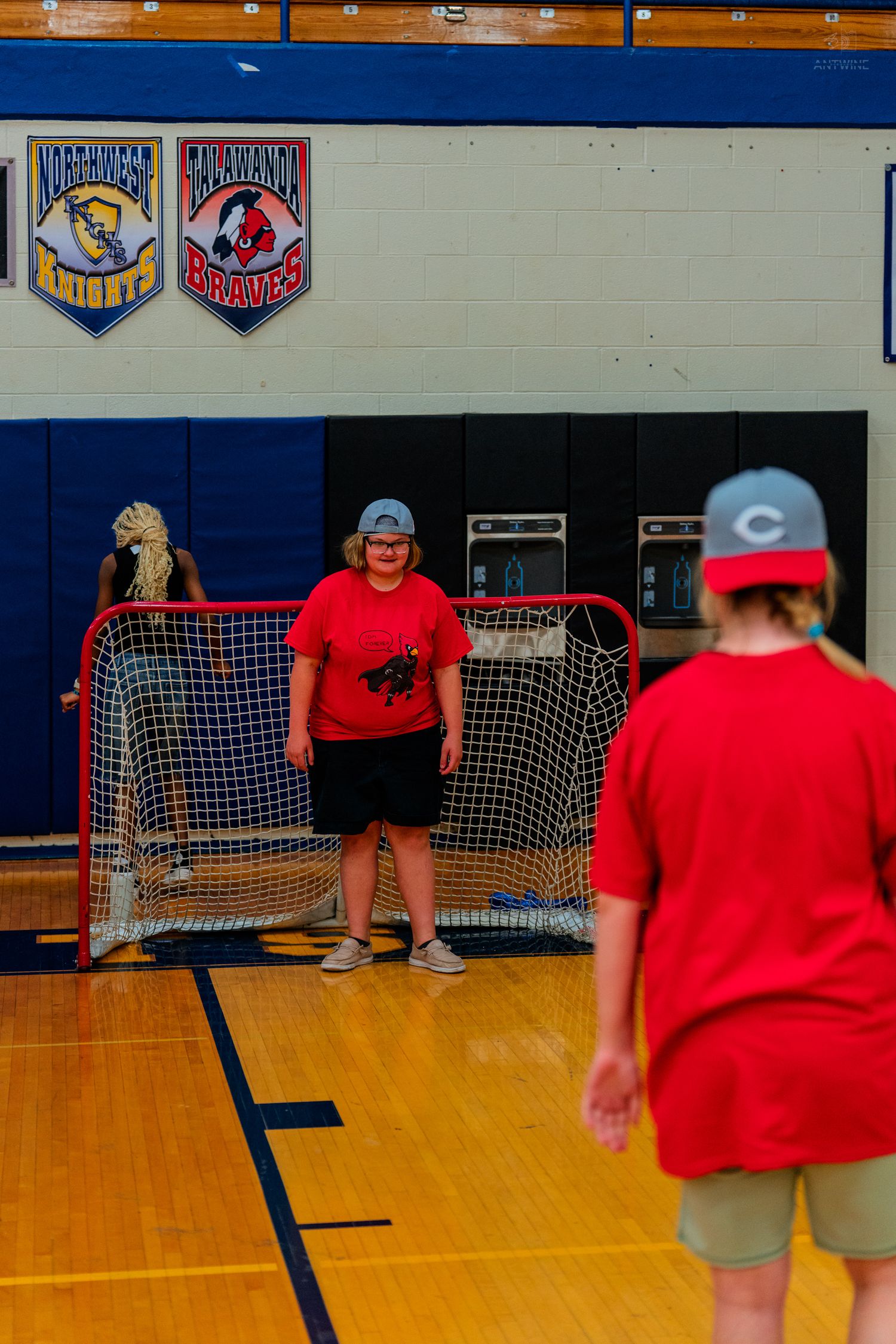 Woman in red shirt stands as goalie in indoor hockey net during gym activity with another player approaching.