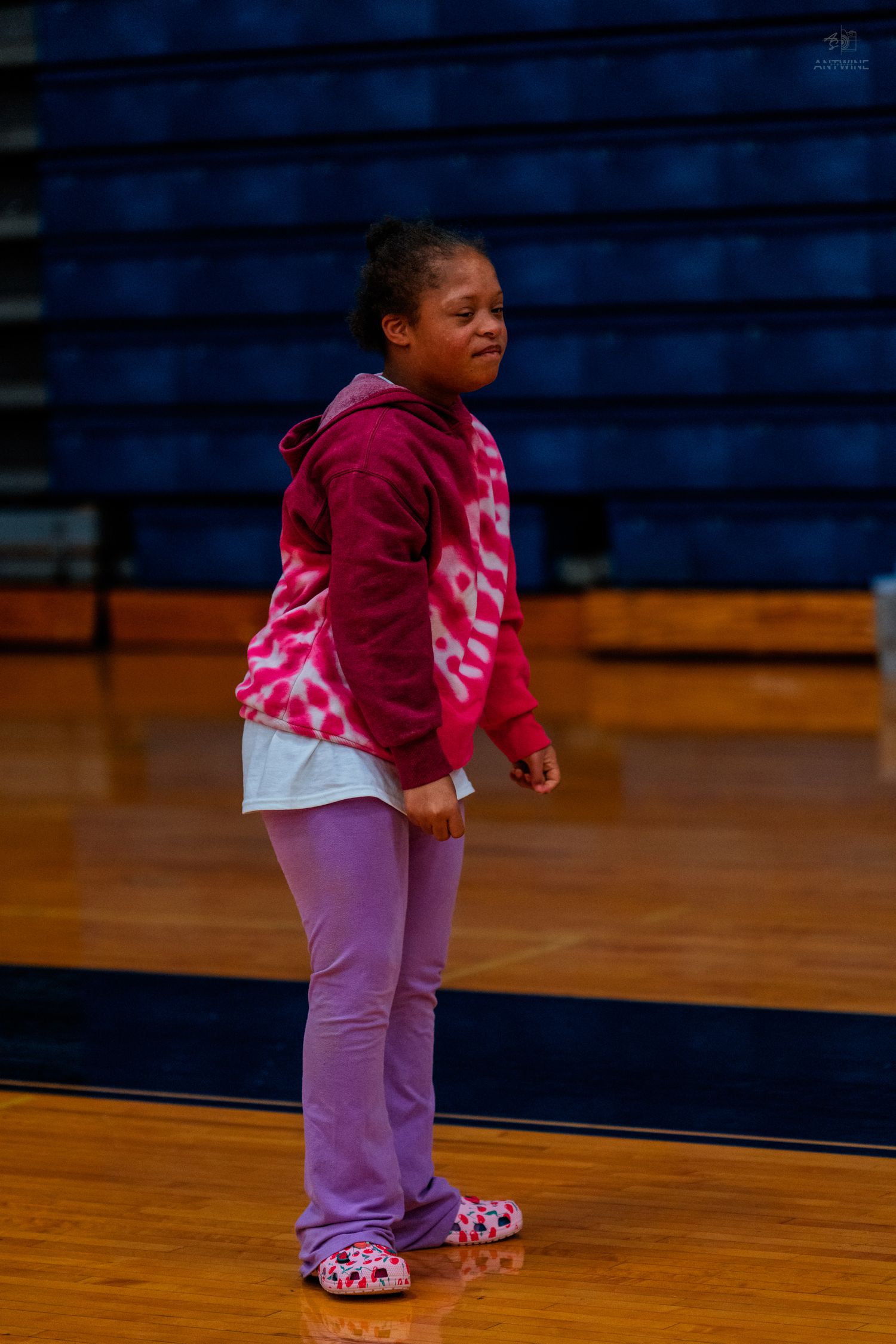 Young girl in pink hoodie and purple pants stands on a gymnasium basketball court floor.