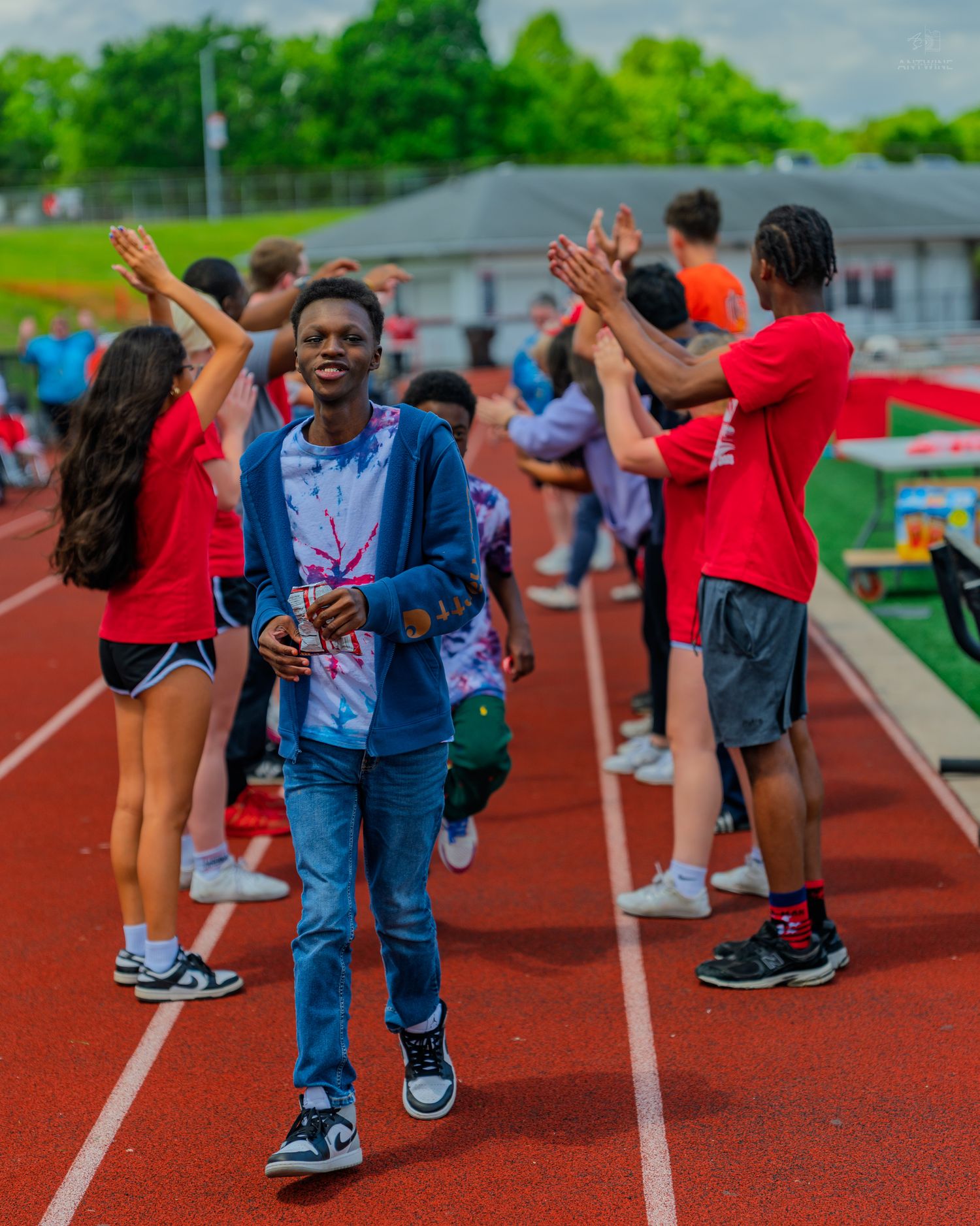 A smiling student walks a red track while peers form a high-five tunnel behind him at a school sports event.