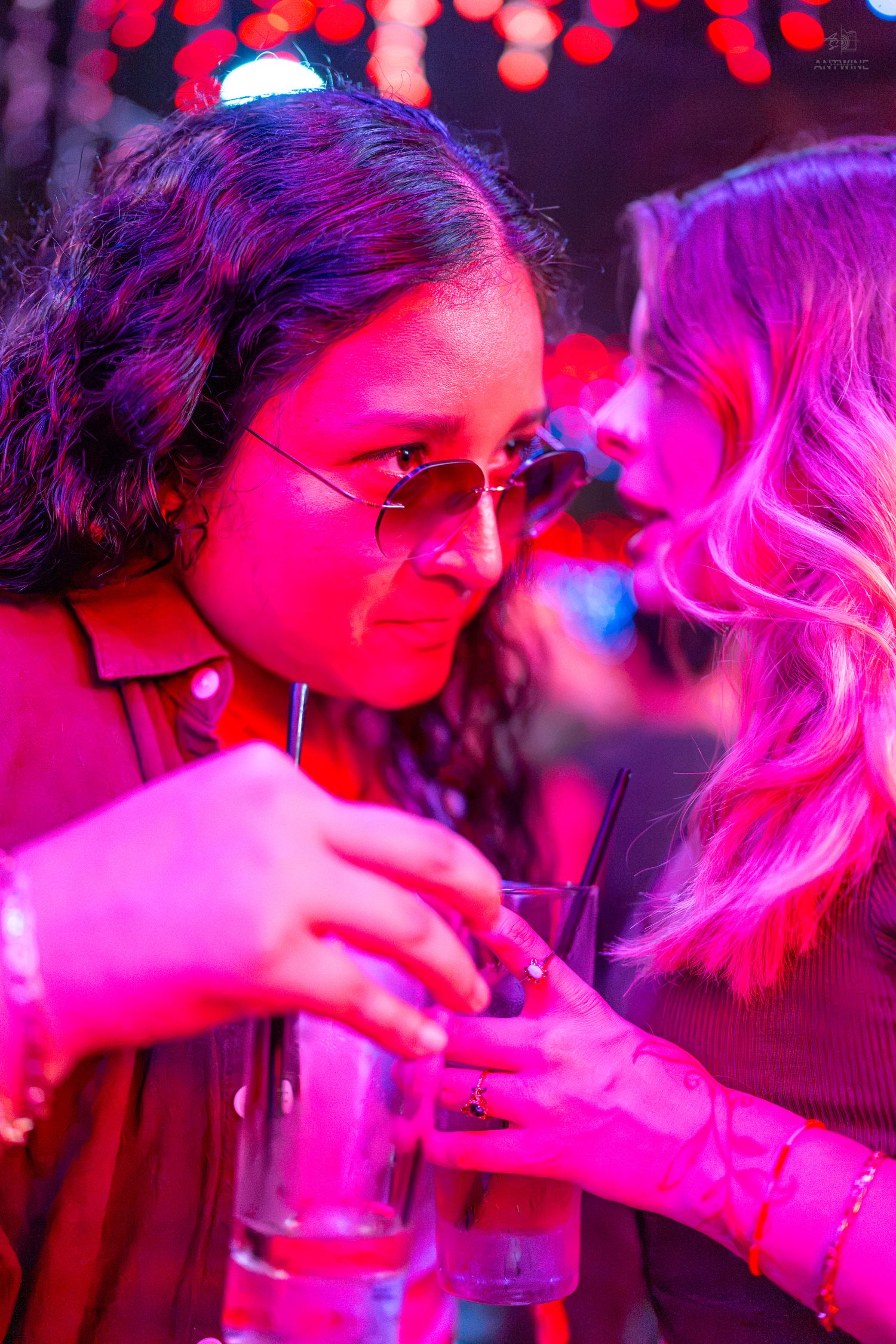 Two women sharing drinks at a vibrant nightclub under pink and red neon lights.