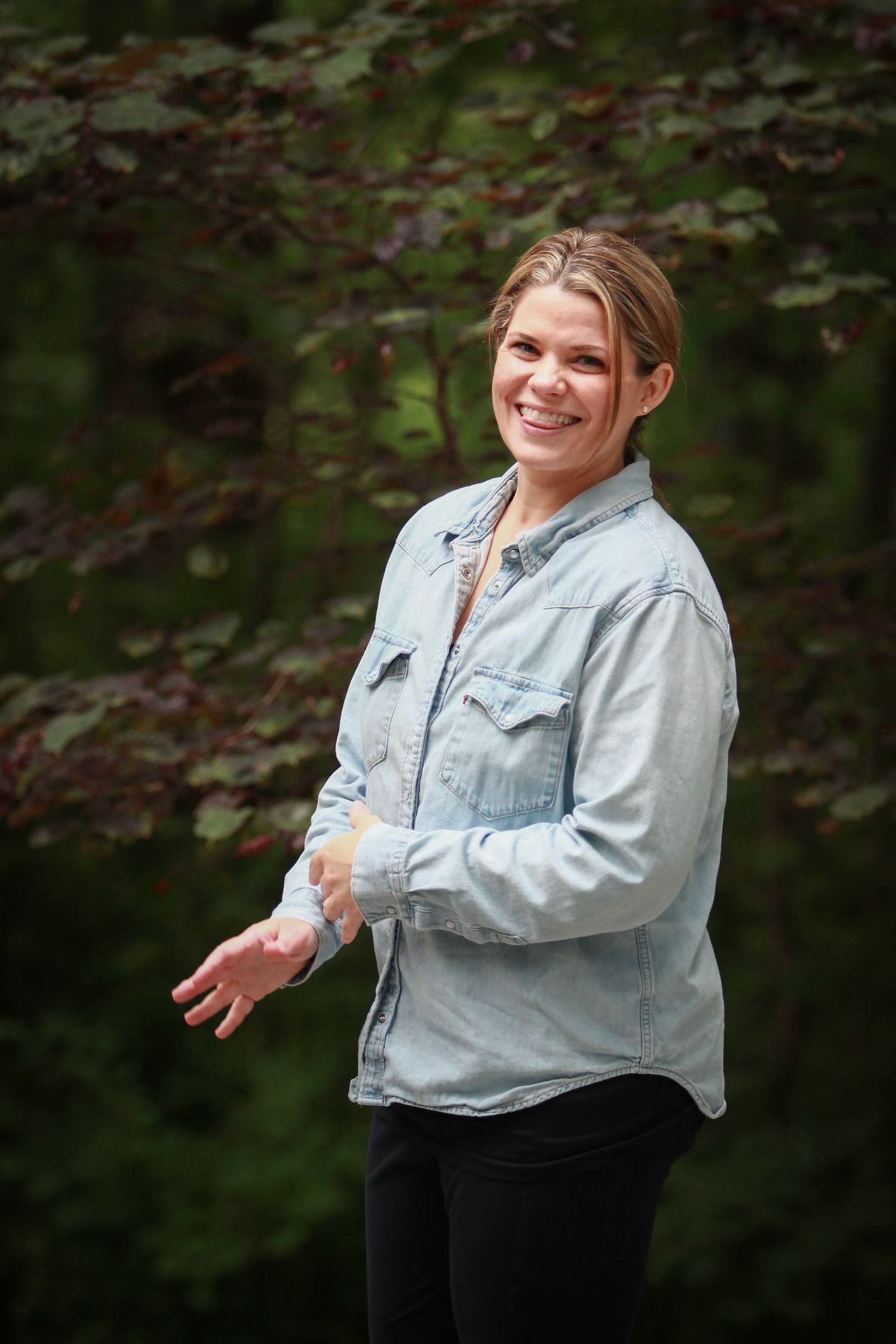 Smiling woman in denim shirt gestures with hands while standing in front of lush green forest background.