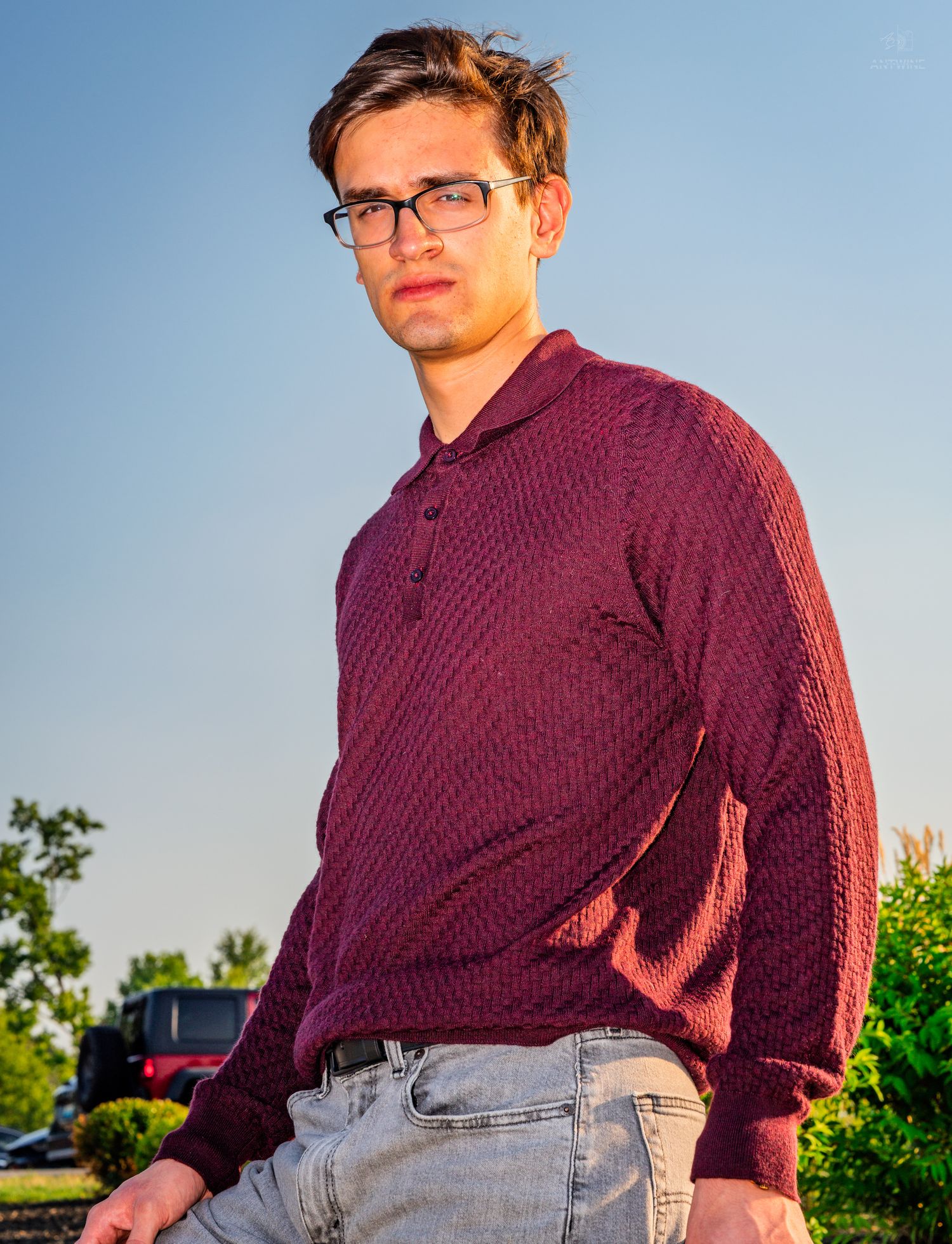 Young man in burgundy knit polo sweater and jeans wearing glasses, posing outdoors on a sunny day.