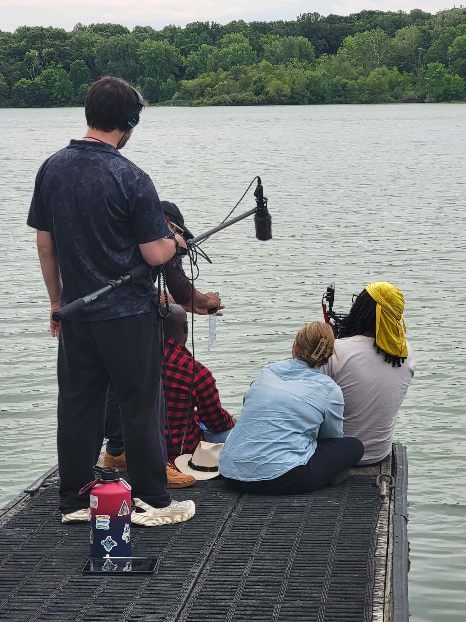 Film crew recording audio and video on a dock overlooking a calm lake surrounded by green trees.